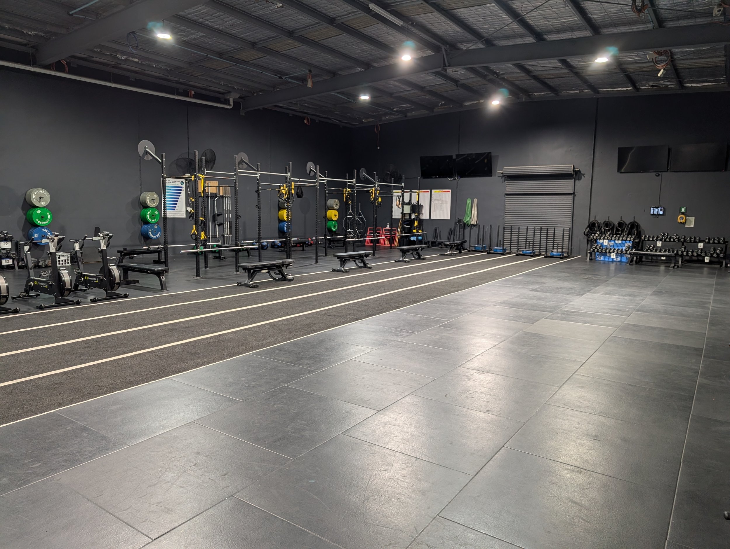 Empty indoor gym with black walls, rubberized flooring, and workout equipment including benches, barbells, weights, and cardio machines.