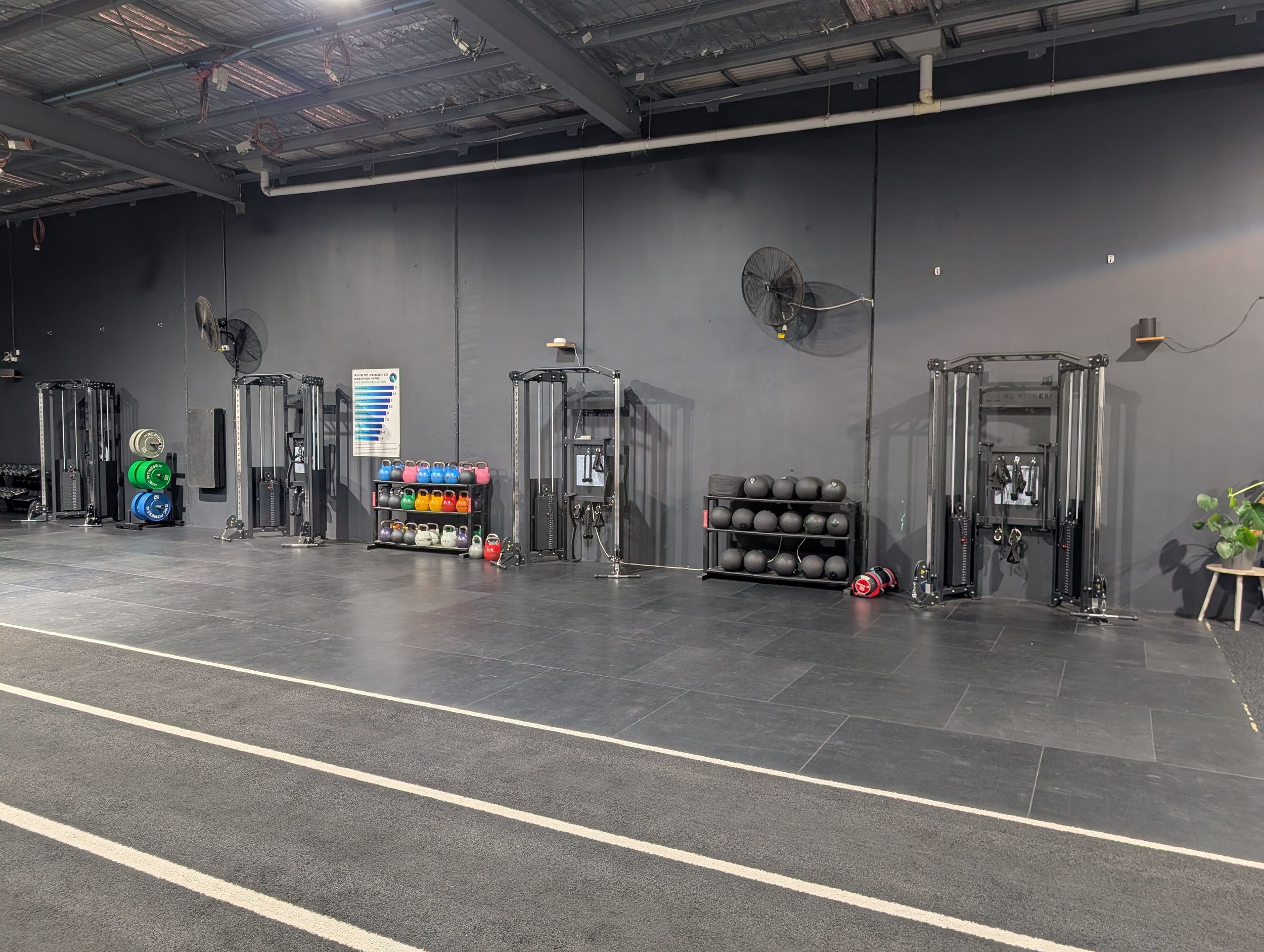 Empty gym with workout equipment including weights, kettlebells, medicine balls, and resistance machines against a dark wall.