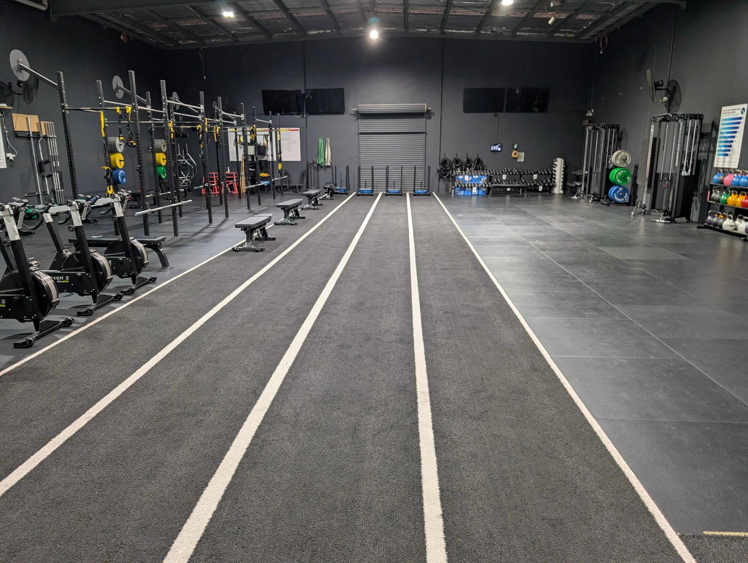 Empty indoor gym with various workout equipment including rowing machines, benches, weights, and racks, with four running lanes on black flooring in the center.