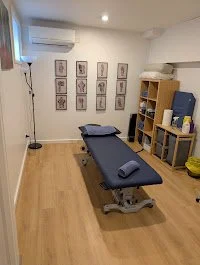 Massage or treatment room with a black massage table, wall art, a tall lamp, and a wooden shelf with towels and supplies.