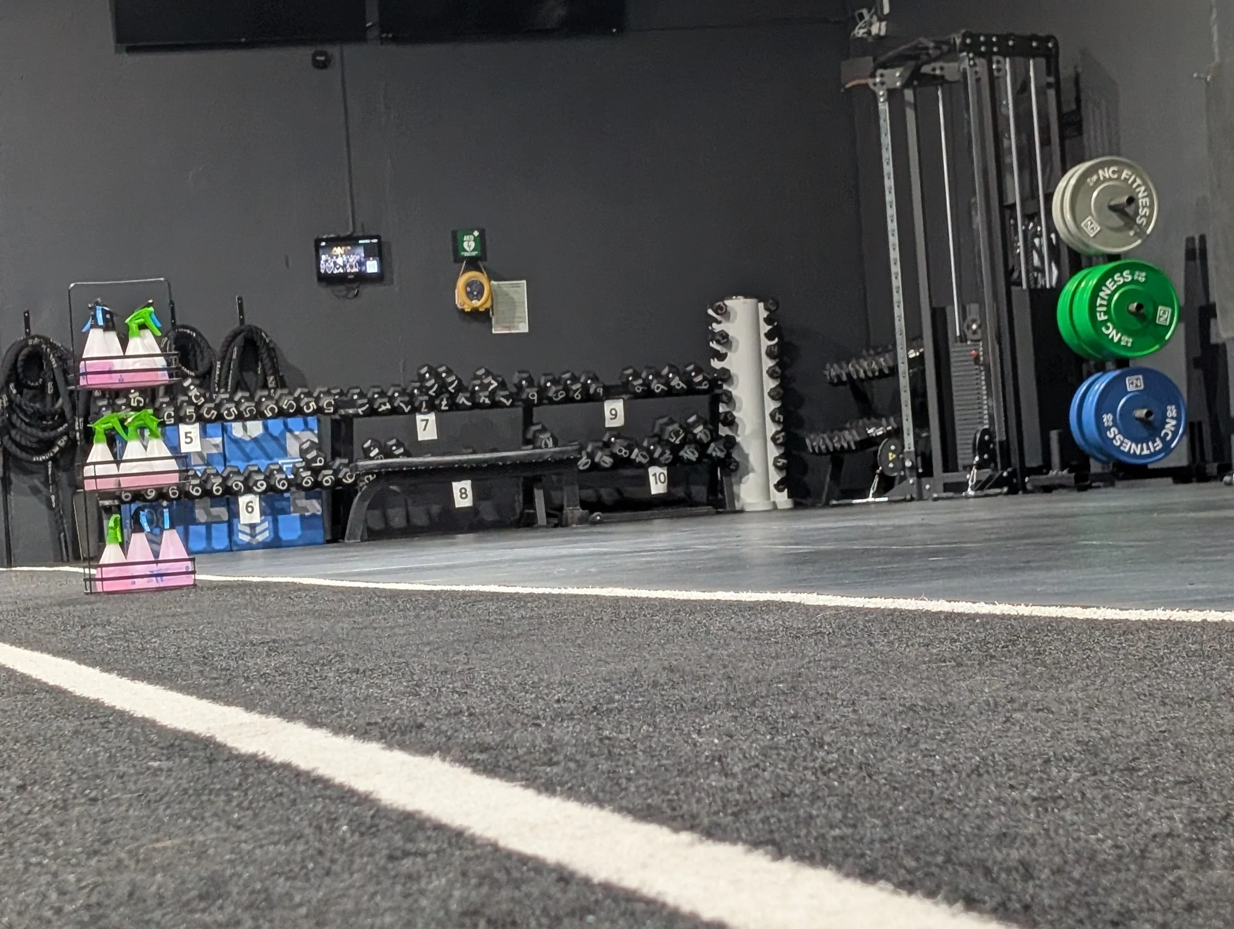 Gym workout equipment including dumbbells, weight plates, resistance bands, and a workout machine, arranged near a black wall with gym accessories in the background.