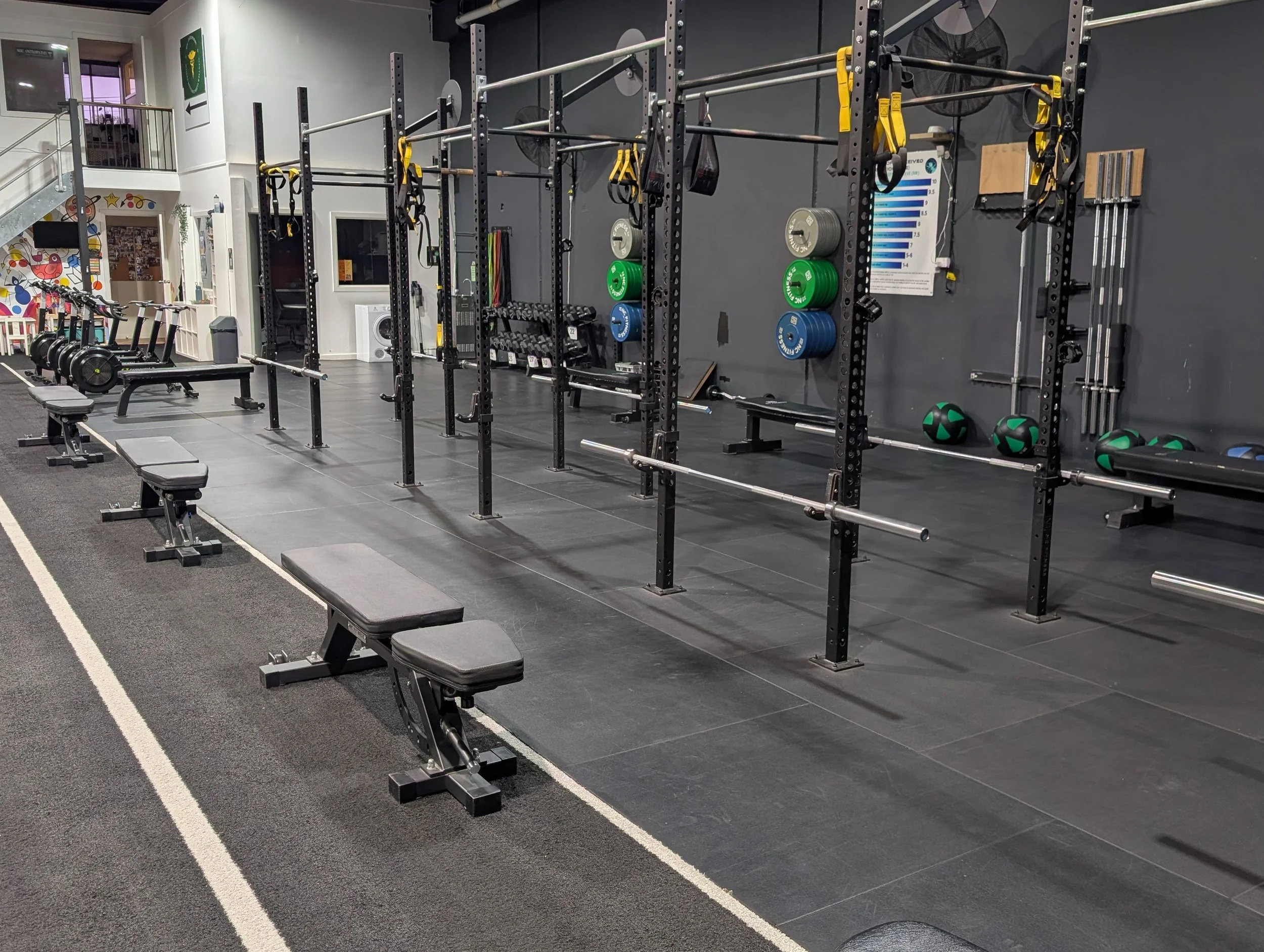Empty gym workout area with black and gray equipment, benches, barbells, kettlebells, weight plates, and resistance bands.