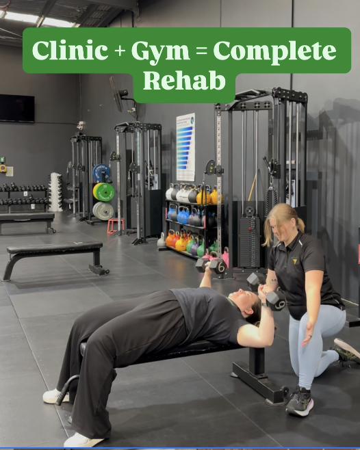 A woman assists a man with a dumbbell exercise on a bench at a gym, with fitness equipment and a motivational sign reading 'Clinic + Gym = Complete Rehab' in the background.