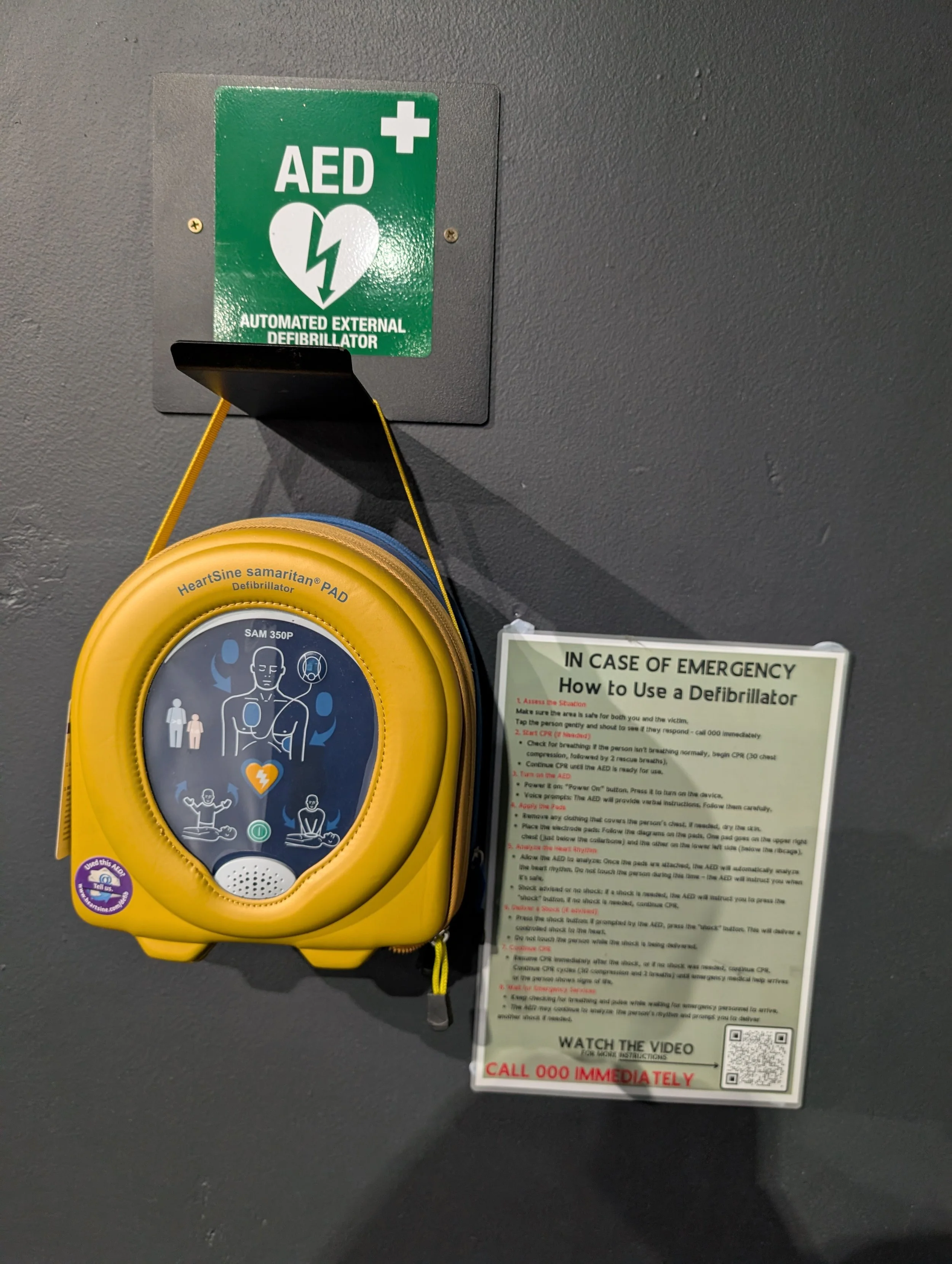 Automated external defibrillator (AED) mounted on a wall with instructions for emergency use and a nearby sign indicating AED location.