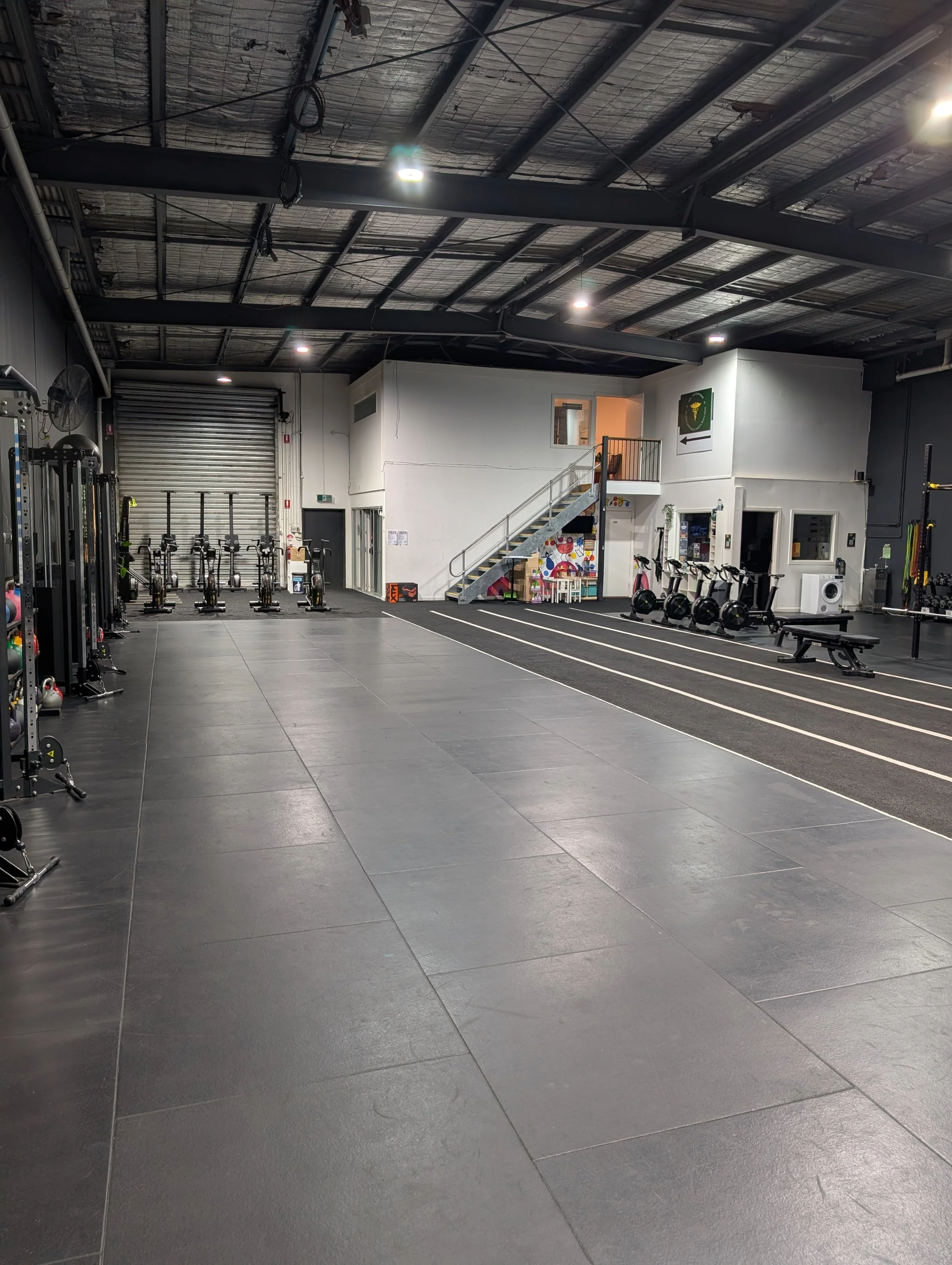 Empty gym with black rubber floor, weightlifting stations on the left, and cardio machines on the right. A staircase leads to a small second level with additional rooms. Gym equipment and a colorful mural are visible in the background.