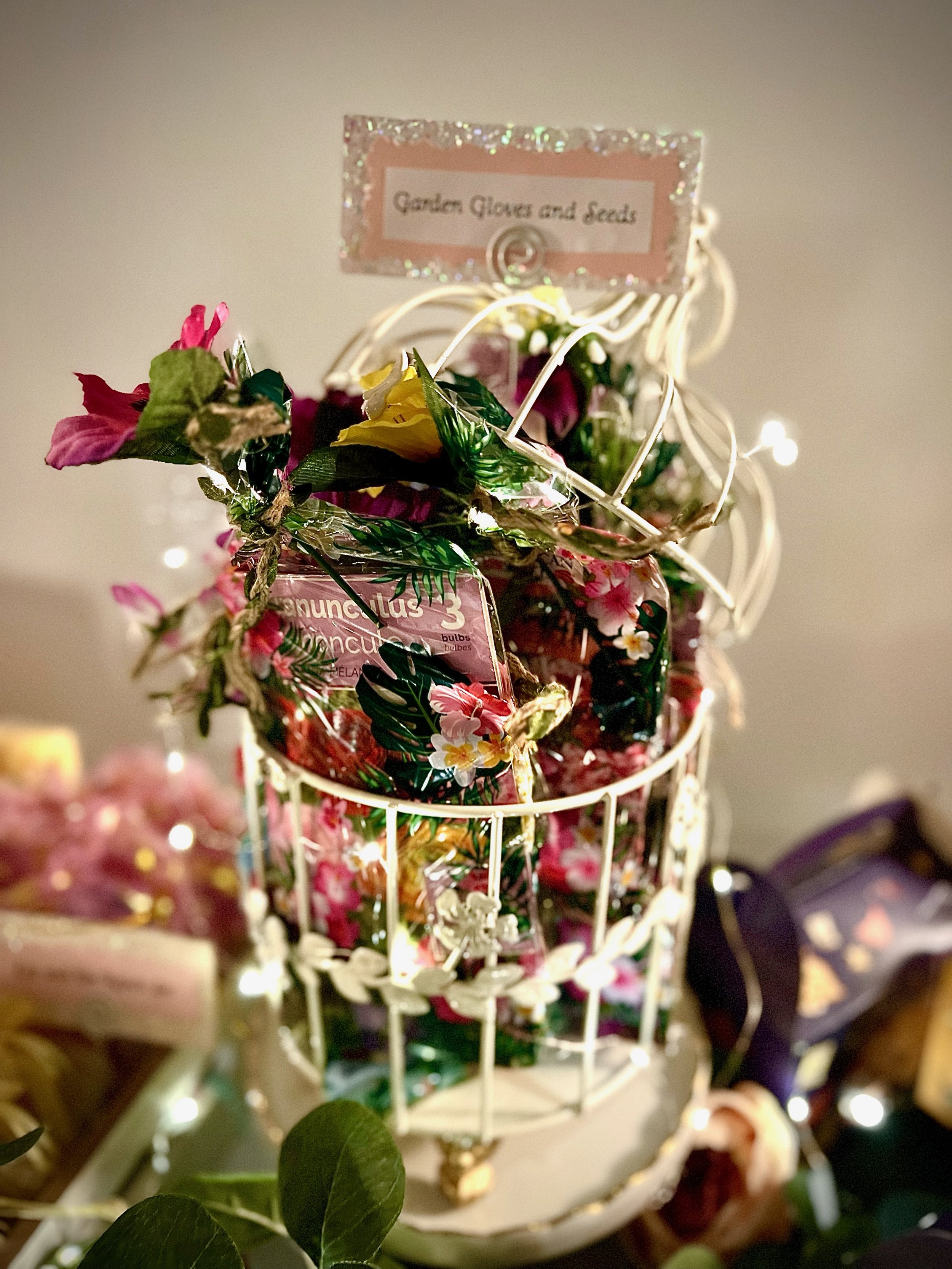 Decorative birdcage filled with colorful artificial flowers, "Garden Gloves and Seeds." The setup is adorned with fairy lights, creating a festive display.