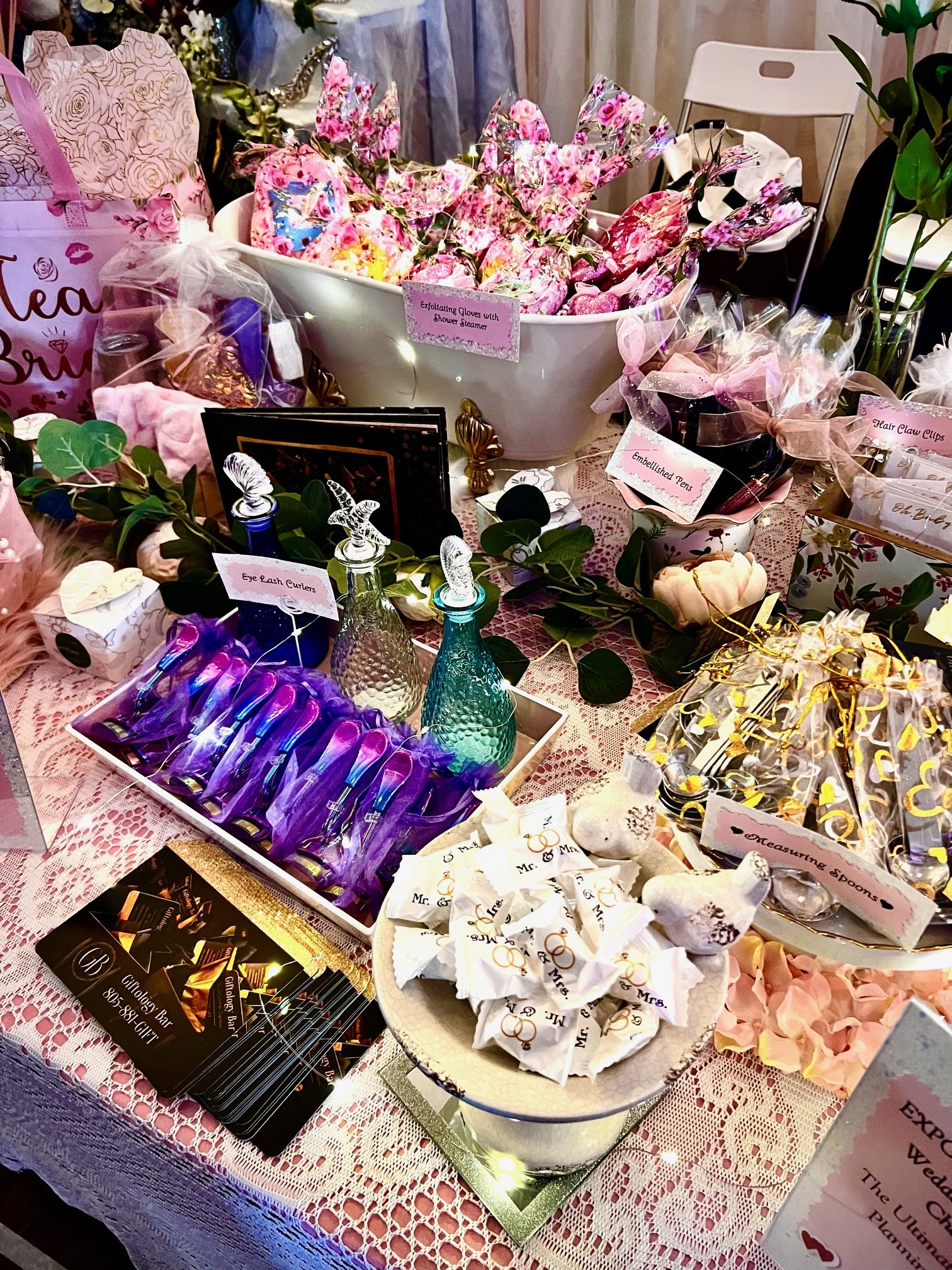 Table display with bridal-themed gifts like exfoliating gloves, measuring spoons, and eye lash curlers, decorated with flowers and lace.