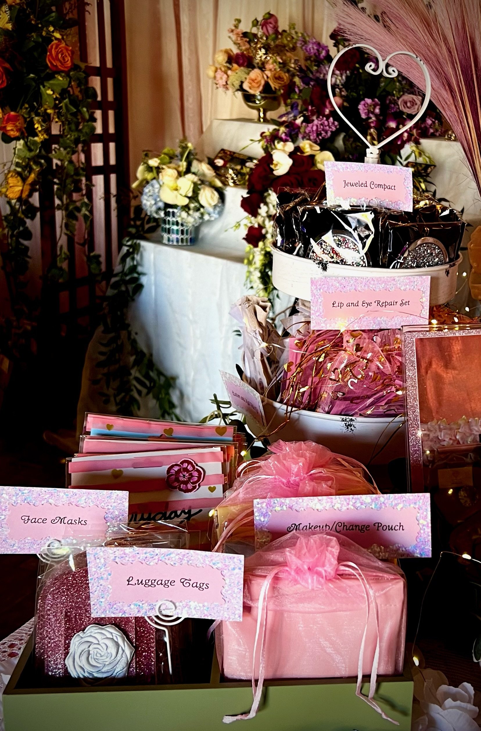 Quinceanera assorted gift items display with pink packaging, including jeweled compacts, lip and eye repair sets, face masks, luggage tags, and makeup pouches, surrounded by floral arrangements.