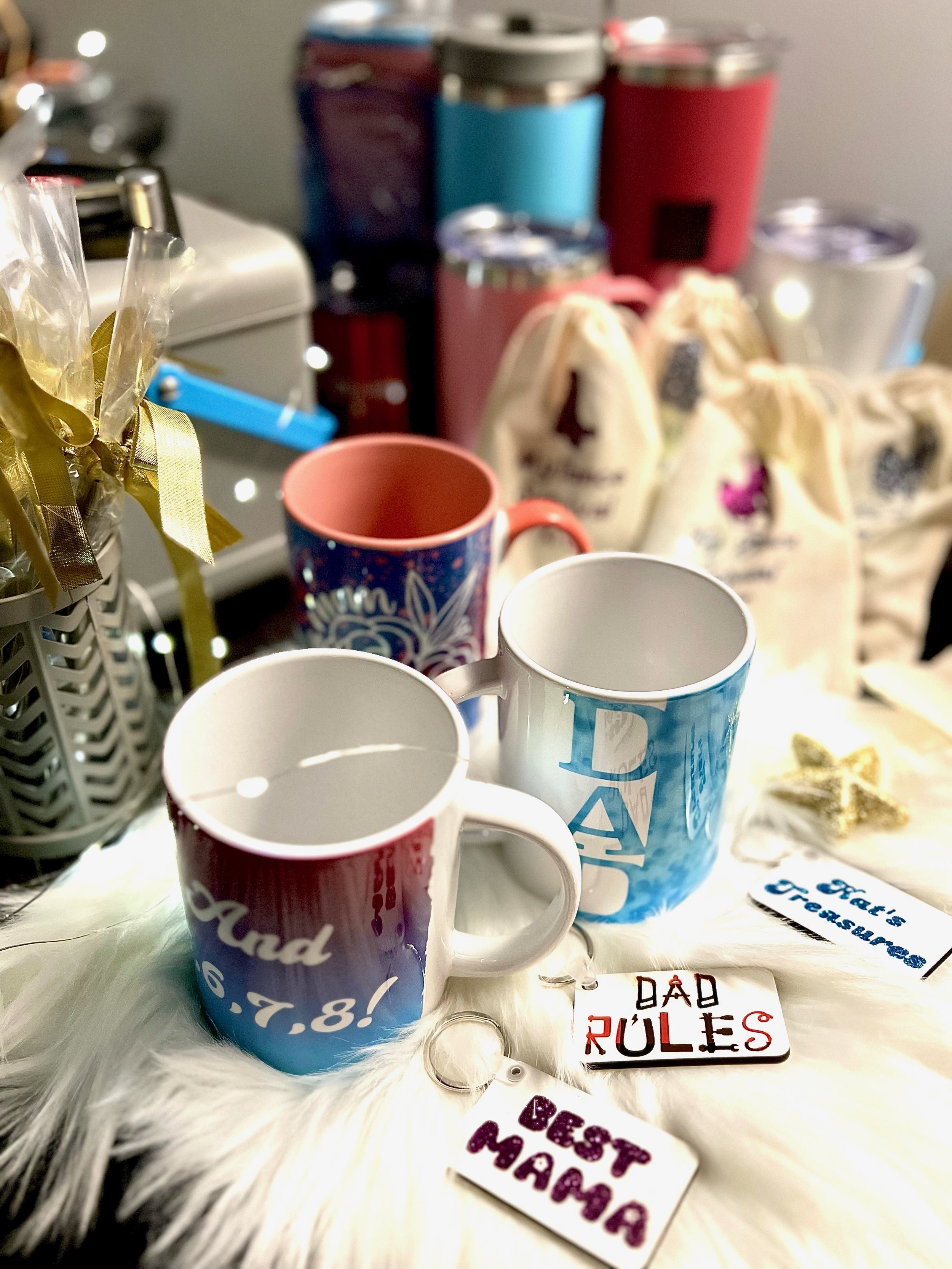 Close-up of mugs with various designs and keychains with text on a white fuzzy surface. Background includes gift bags and tumblers.