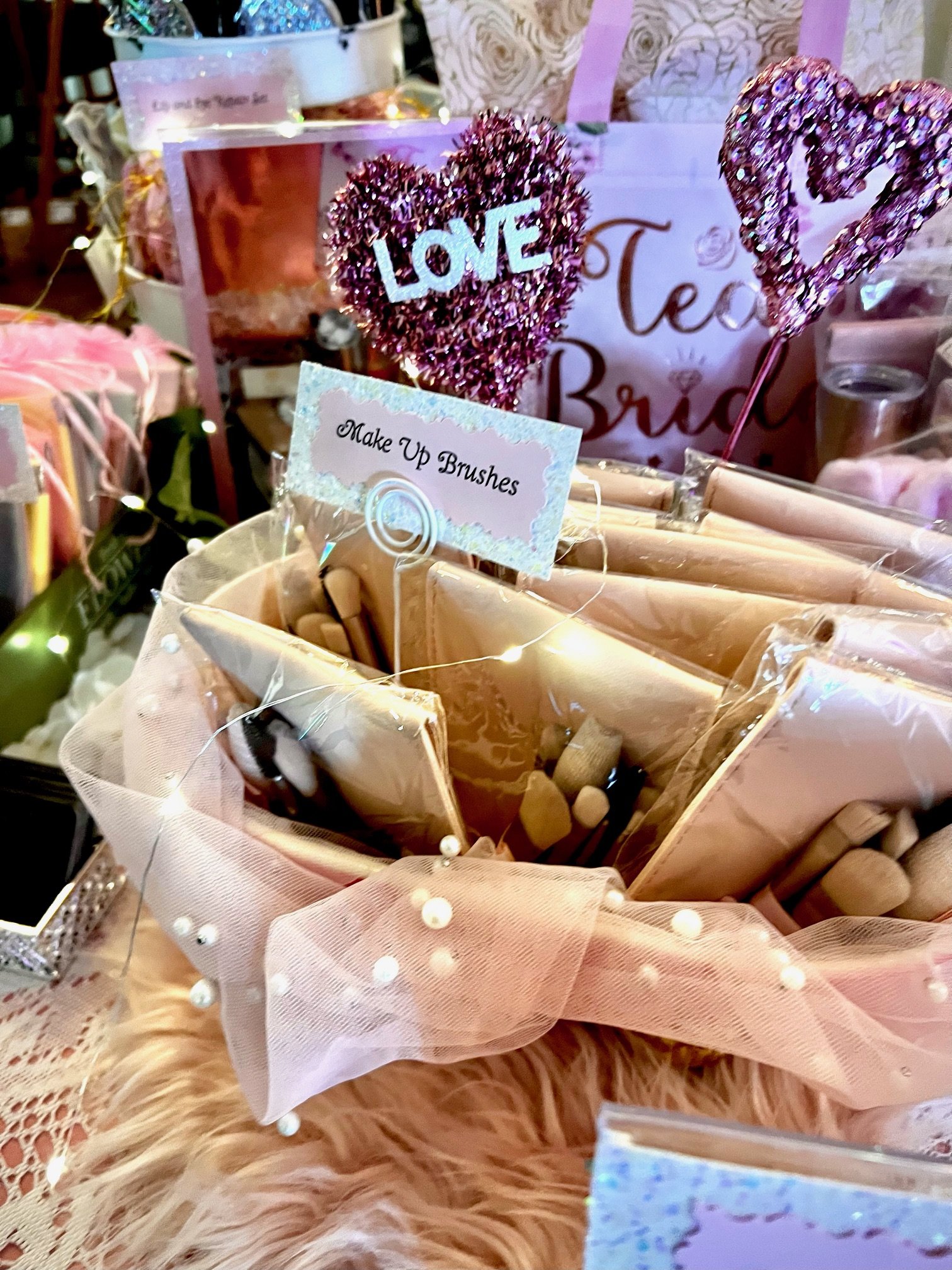 Display of makeup brushes in a decorative arrangement. The setup includes soft fabrics, fairy lights, and a sign labeled "Make Up Brushes," with a bridal theme in the background.