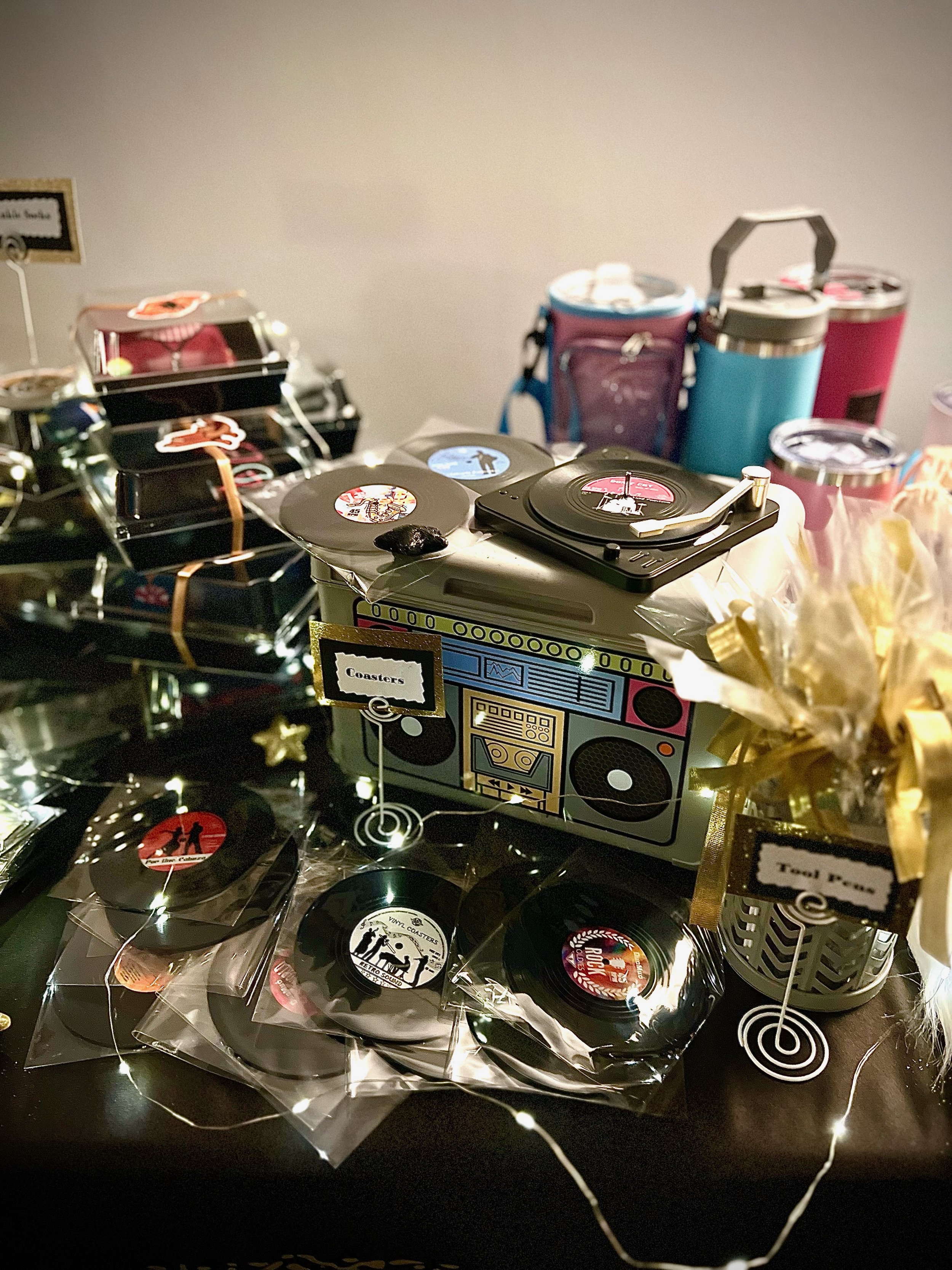 A decorative display featuring a retro-themed setup with vinyl record coasters in clear sleeves, and colorful insulated drink tumblers. The scene is adorned with string lights and gift-wrapped items, creating a festive atmosphere.