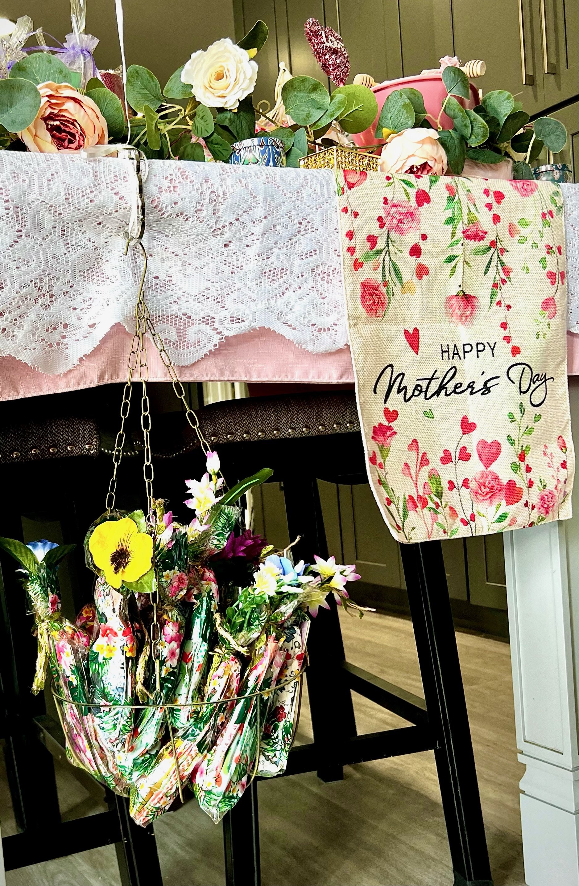 Mother's Day decorations with floral arrangement, lace cloth, and a hanging basket with wrapped seeds and gloves.