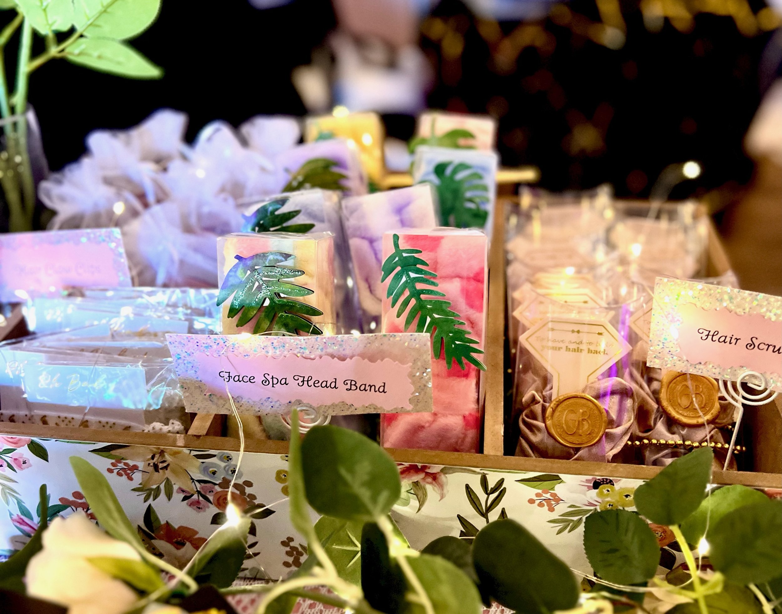Display of spa and beauty products including face spa head bands, bath bombs, hair scrunchies, and soap with decorative packaging. Floral and greenery accents.