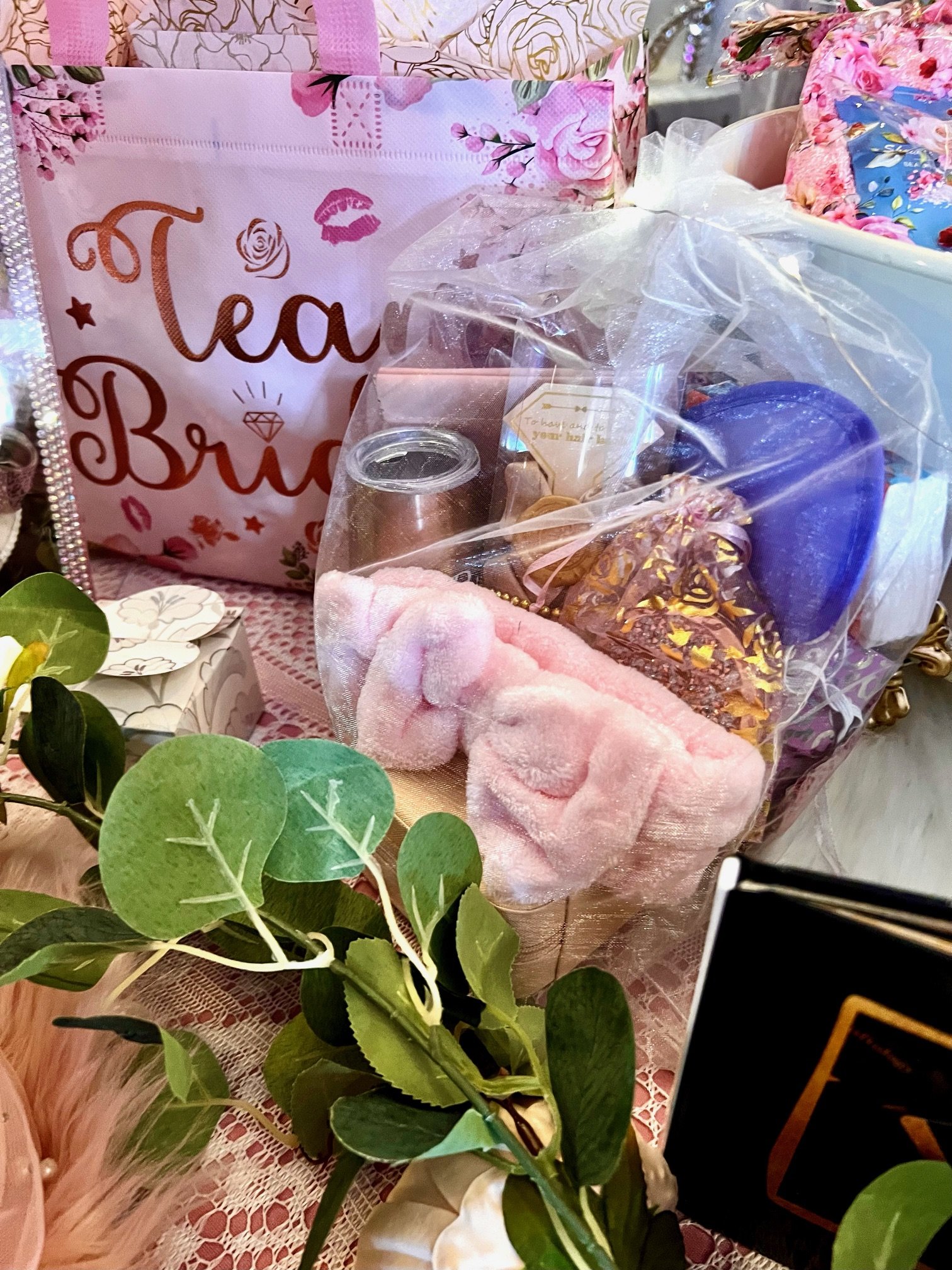 Gift bag labeled "Team Bride" surrounded by pink and floral packaging, including a transparent bag with spa items like a scrub, lotion, and slippers, placed on a decorated table with greenery.