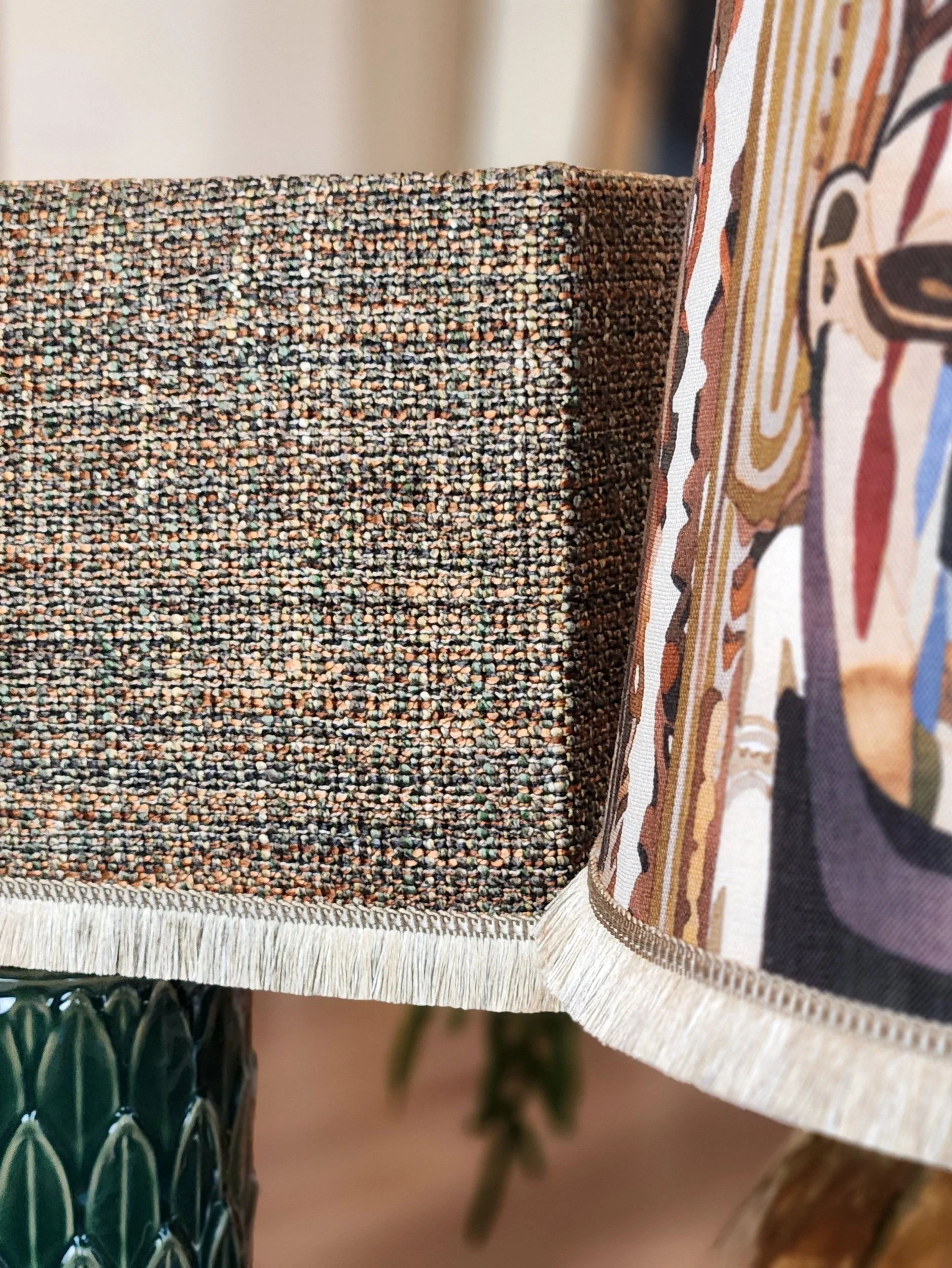 Close-up of two different patterned fabric lampshades, one with a multicolored, tightly woven texture and the other with a colorful, abstract printed design, with fringed edges.