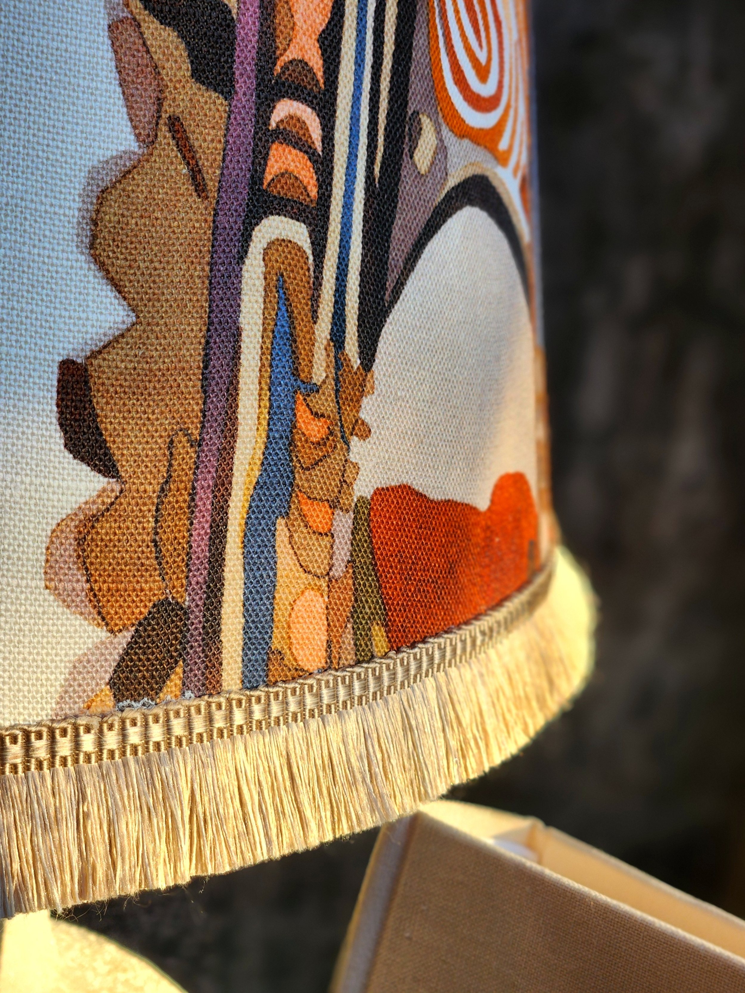 Close-up of a decorative lampshade with a patterned fabric featuring abstract and floral designs, fringed trim, and a yellow base.