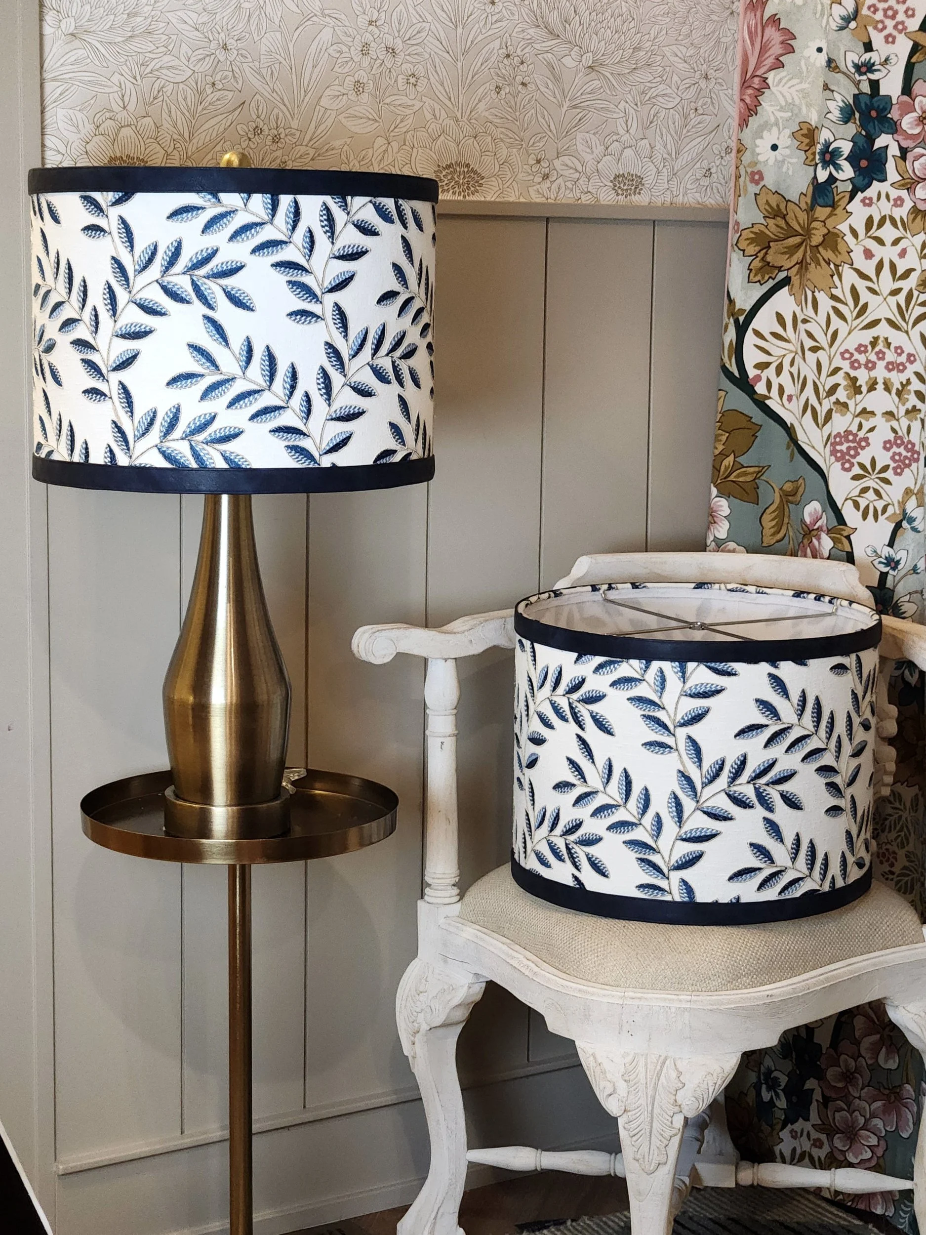 A gold-colored table lamp with a leaf-patterned lampshade, and a matching leaf-patterned lampshade placed on a white vintage-style wooden chair in front of floral wallpaper.