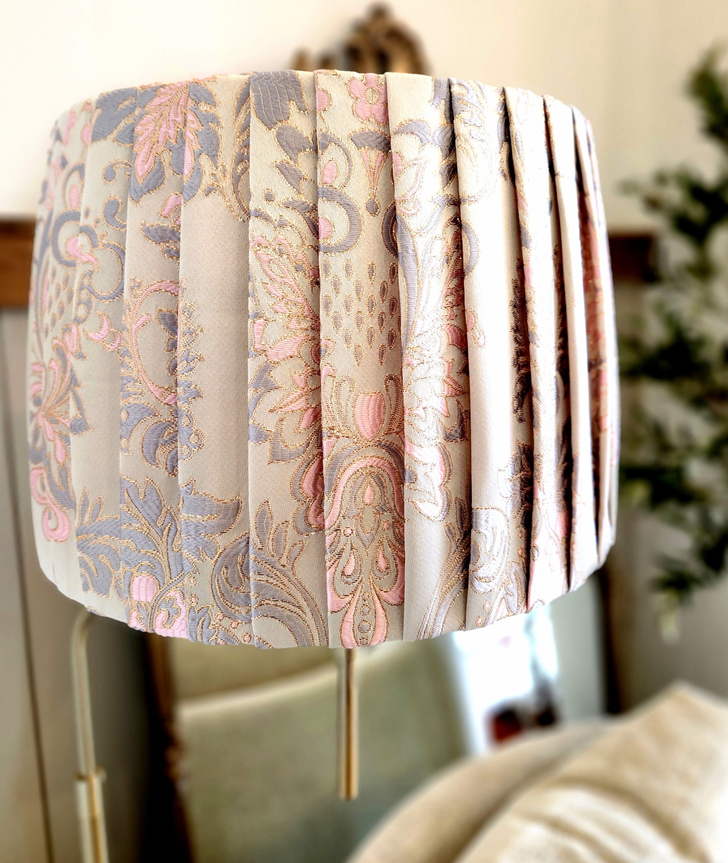 A lampshade with a cream fabric featuring a pink and grey metallic damask pattern. The fabric is pleated to create a textured effect.