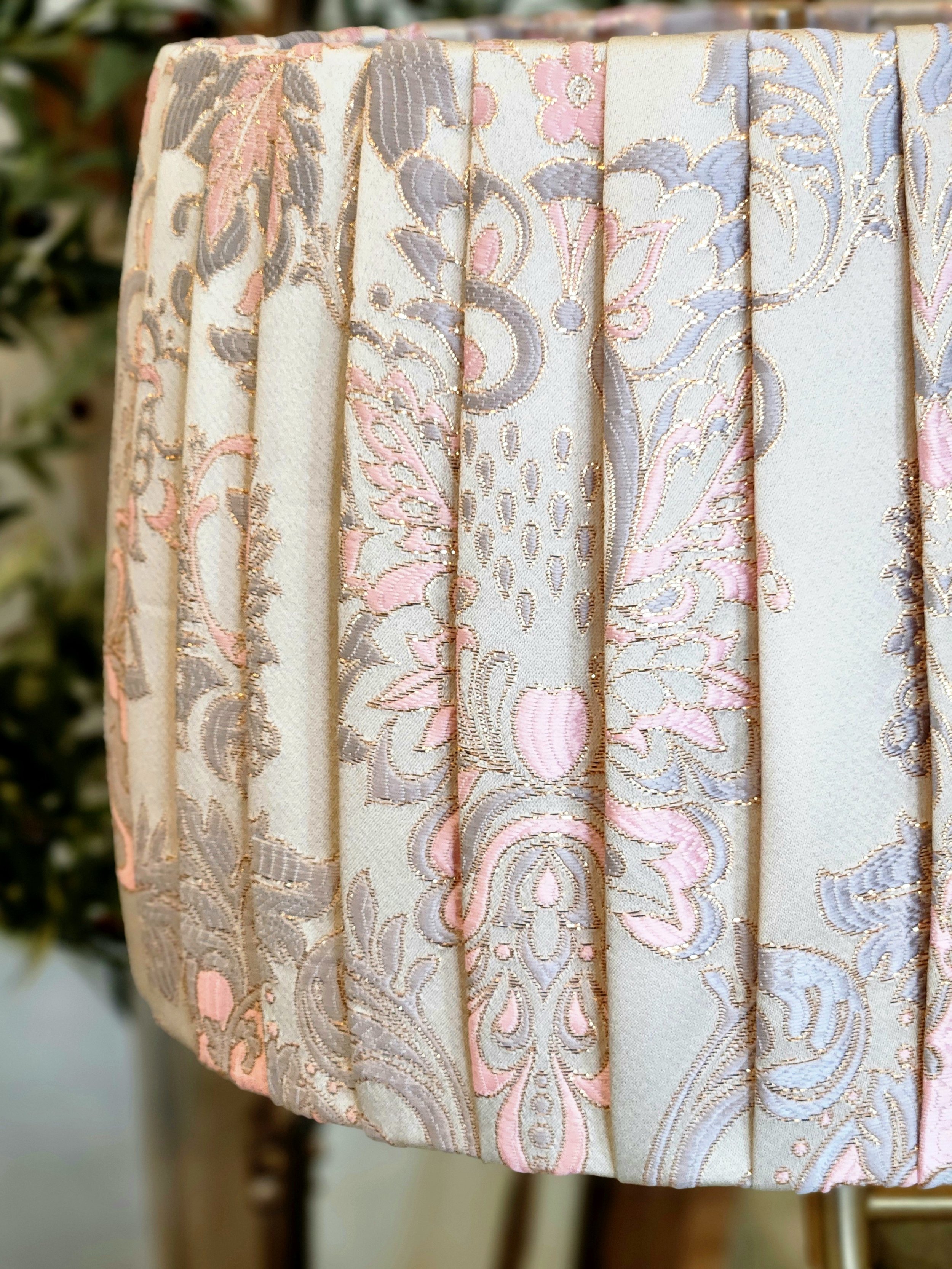 Close-up of a pleated lampshade with a cream fabric and a pink, blue, and gold floral damask pattern. The pleats add texture and dimension to the shade.