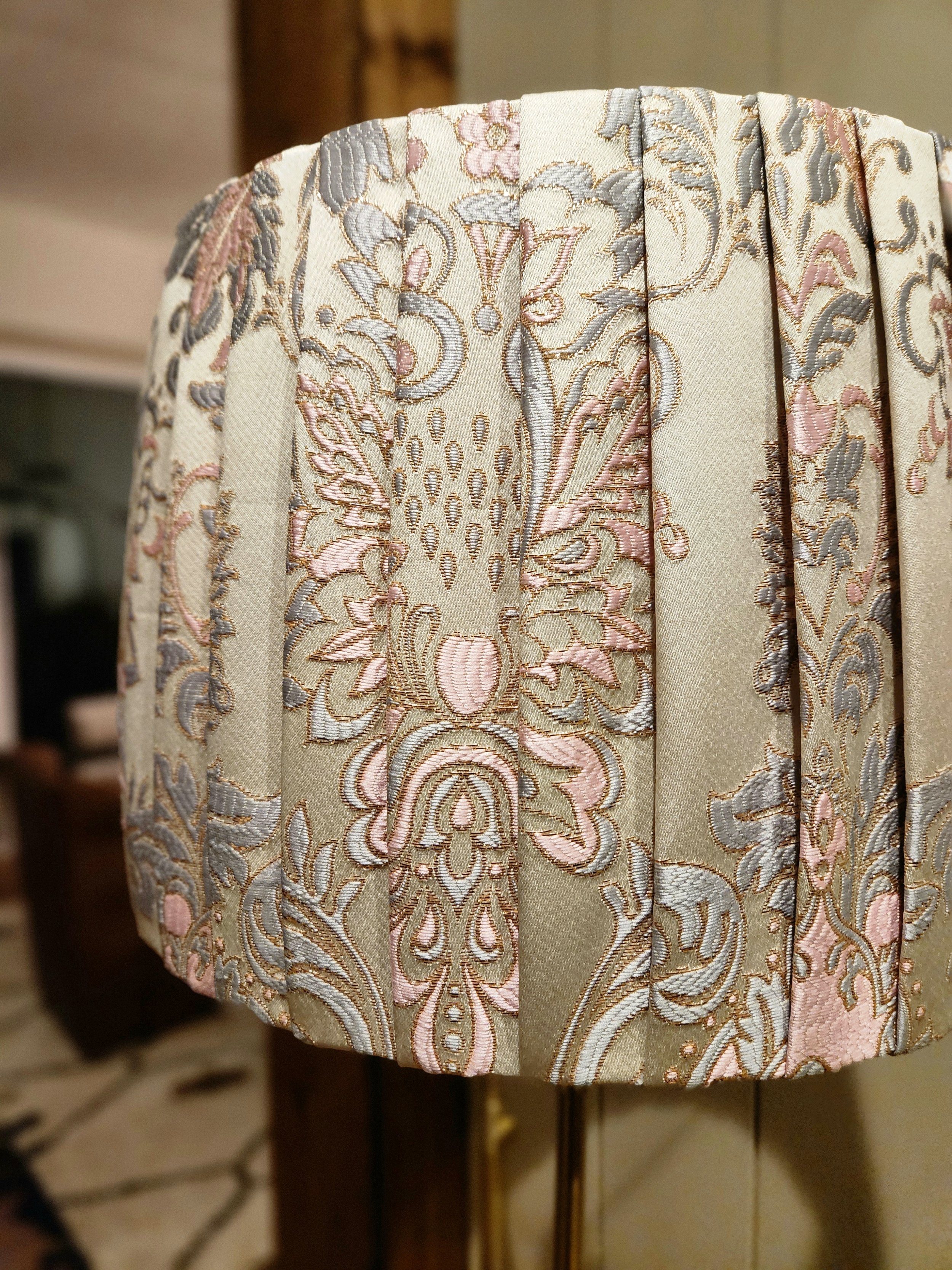 Close-up of a pleated lampshade with a cream fabric and a pink, blue, and gold floral damask pattern. The pleats add texture and dimension to the shade.