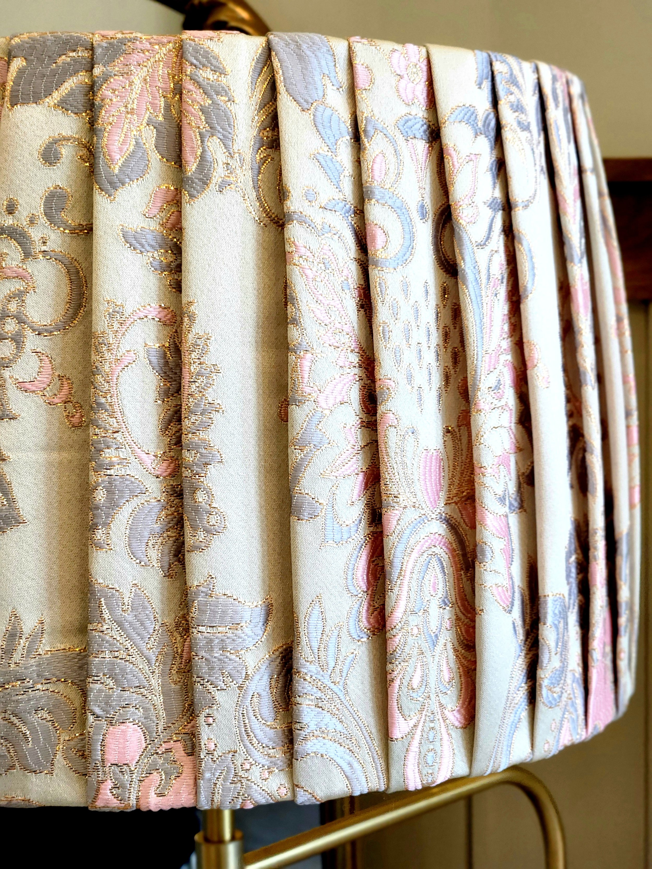 Close-up of a pleated lampshade with a cream fabric and a pink, blue, and gold floral damask pattern. The pleats add texture and dimension to the shade.