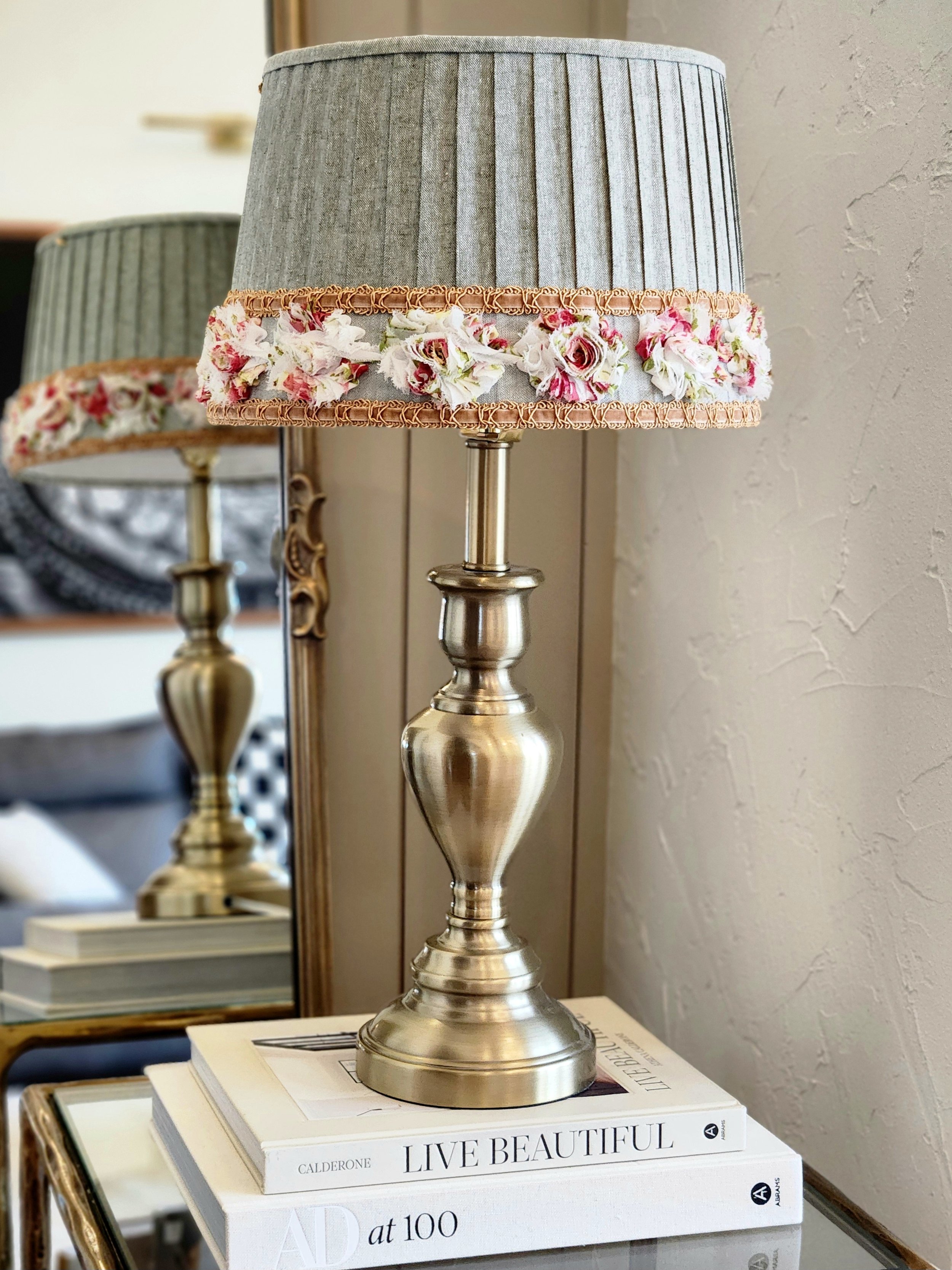 A stylish table lamp with a sage green pleated shade, accented with a floral ribbon and gold braid trim. The lamp sits on a stack of books next to a mirror.
