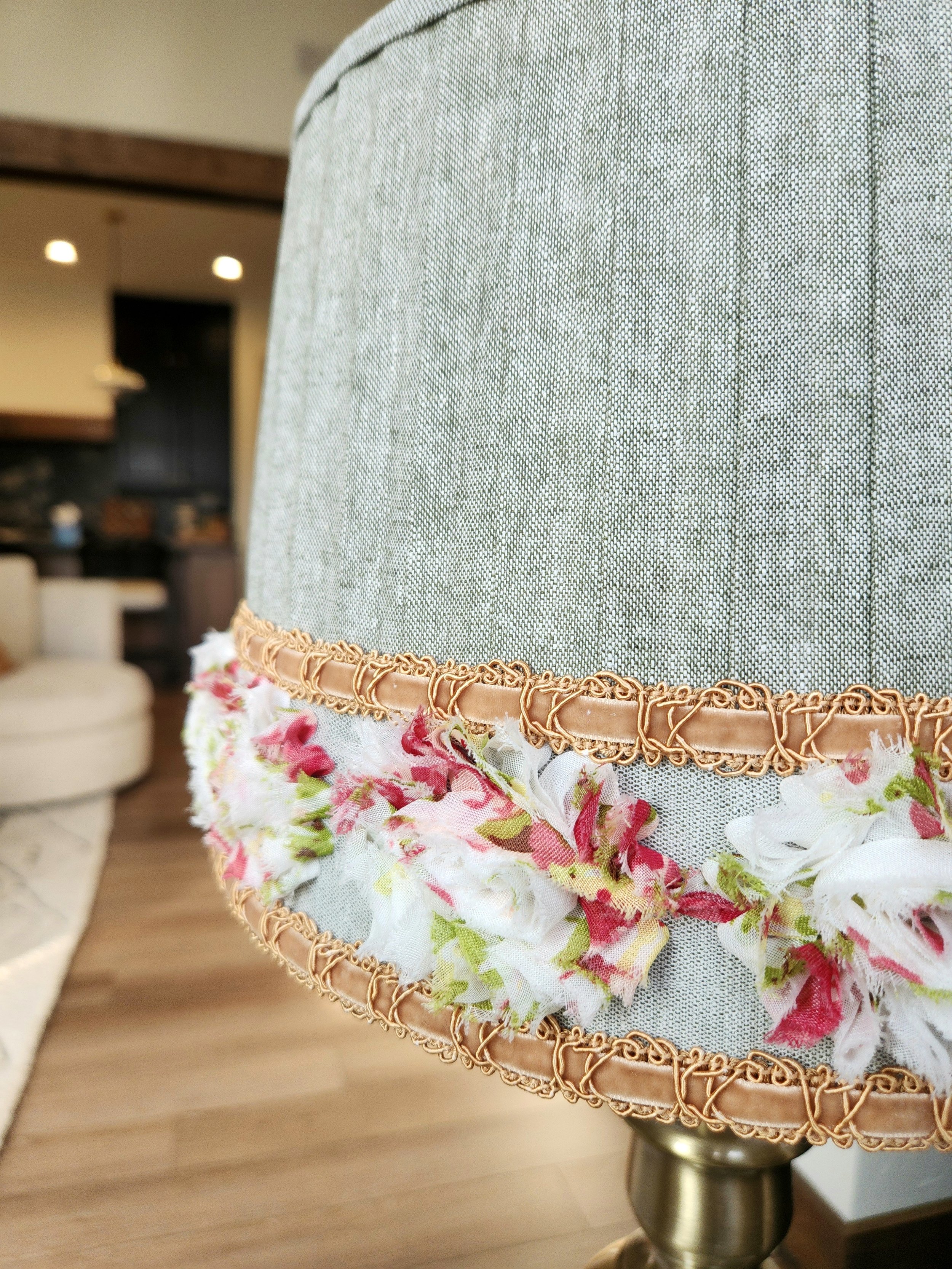 Close-up of a sage green pleated lampshade. It's decorated with a layered trim featuring a gold braided cord and a floral ribbon with red, white, and pink flower accents.
