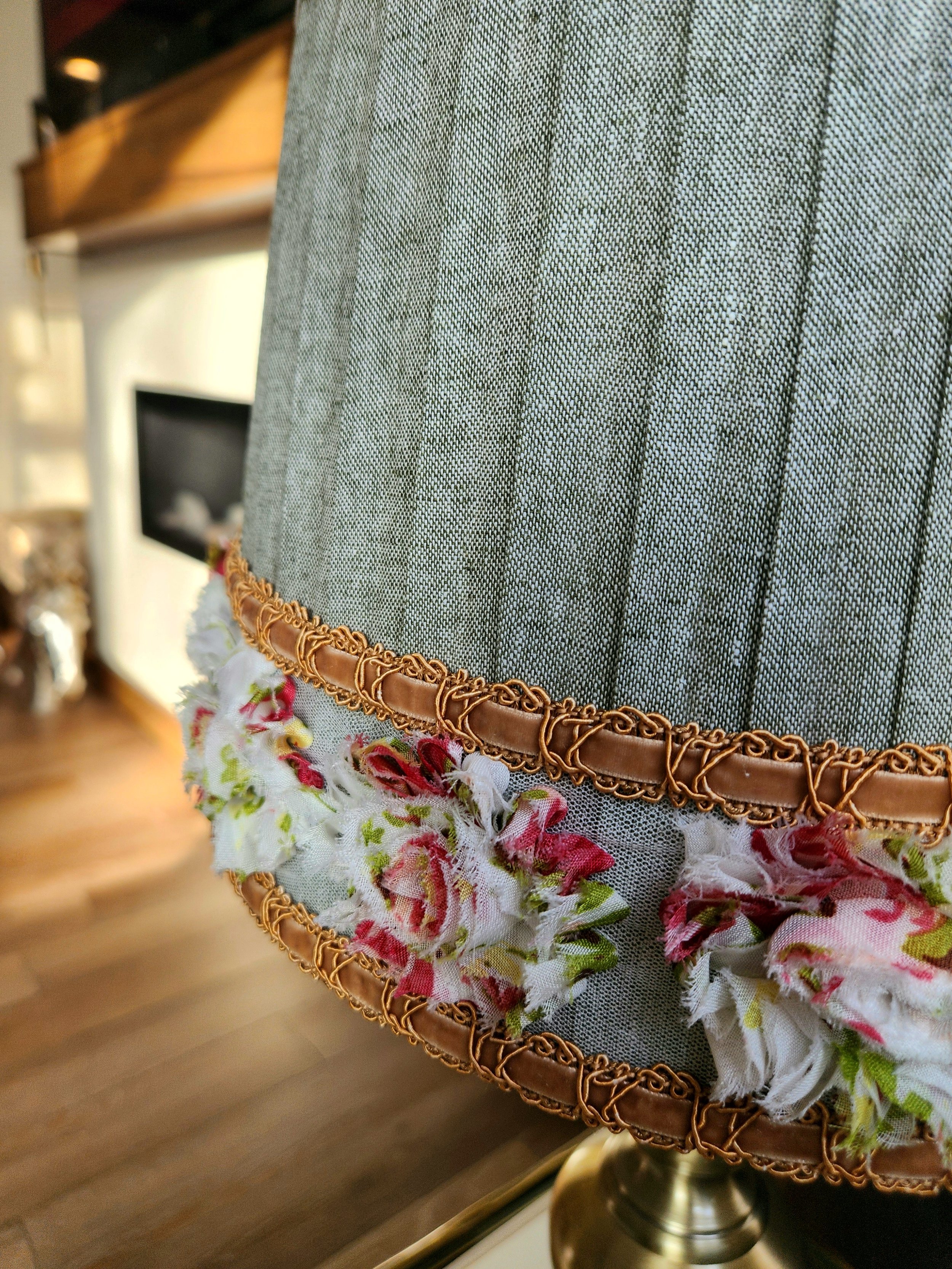 Close-up of a sage green pleated lampshade. It's decorated with a layered trim featuring a gold braided cord and a floral ribbon with red, white, and pink flower accents.