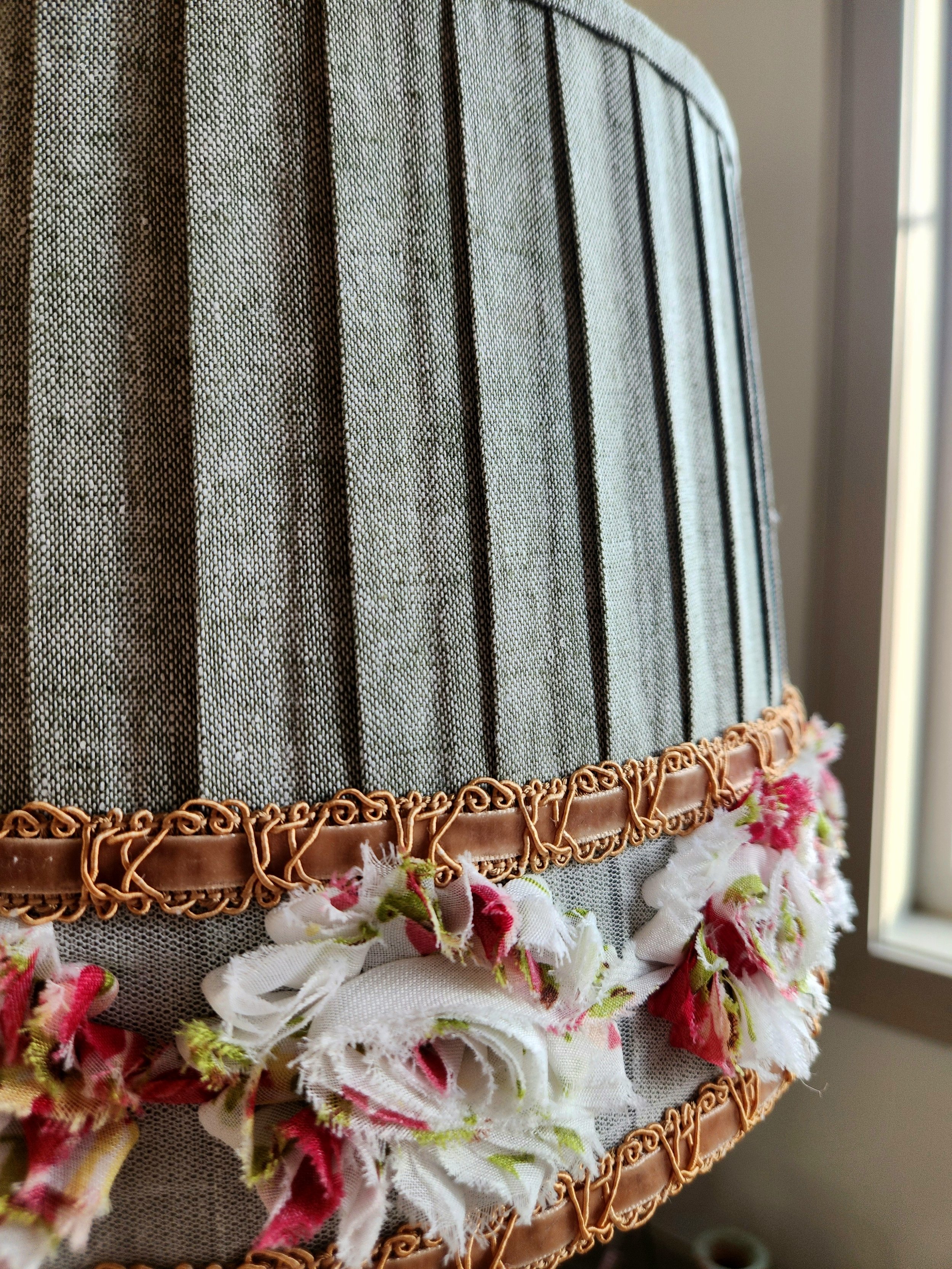 Close-up of a sage green pleated lampshade. It's decorated with a layered trim featuring a gold braided cord and a floral ribbon with red, white, and pink flower accents.