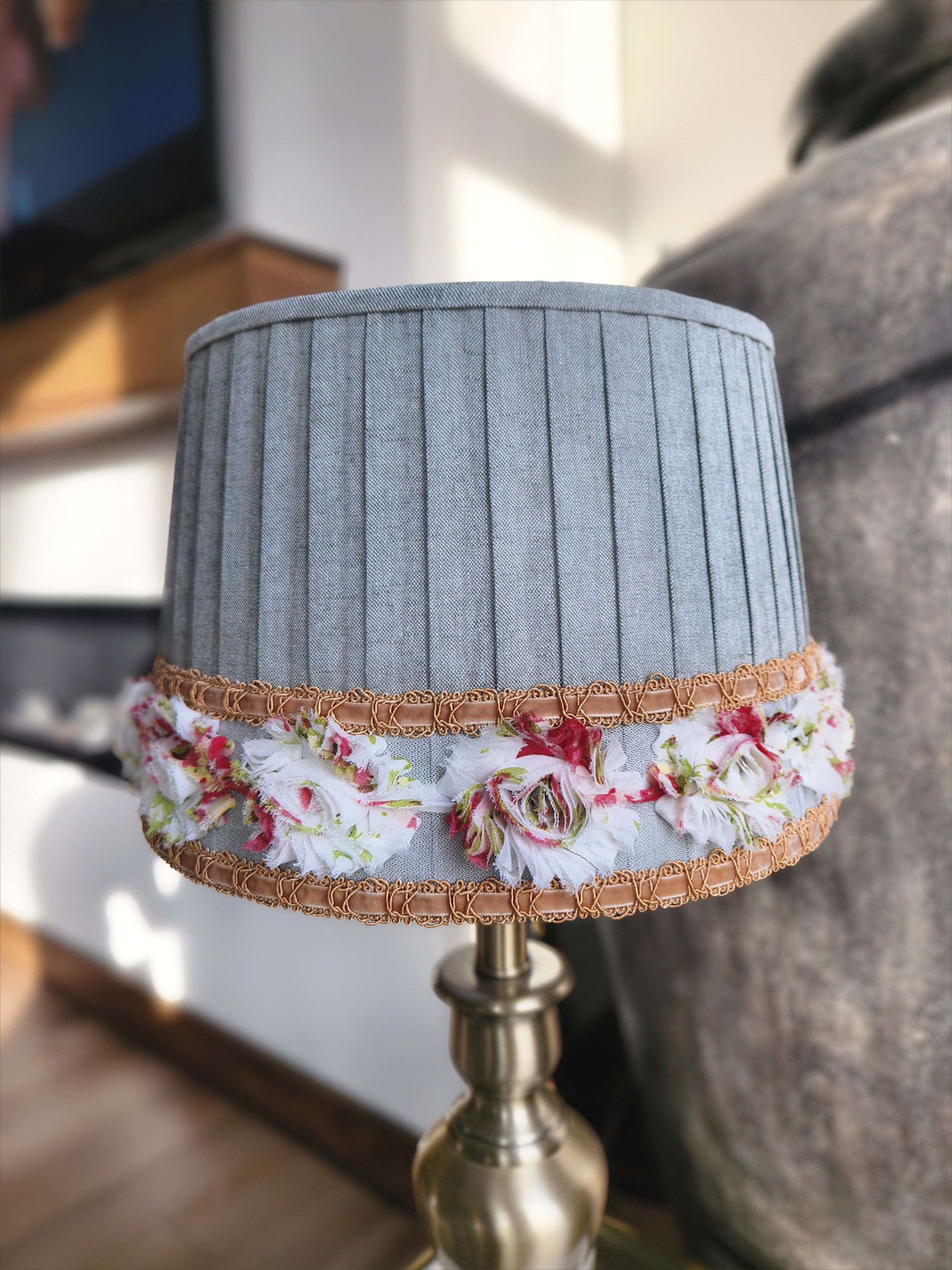 A sage green pleated lampshade adorned with a delicate floral ribbon trim. The ribbon features red, pink and white flowers and is bordered by a gold braid.