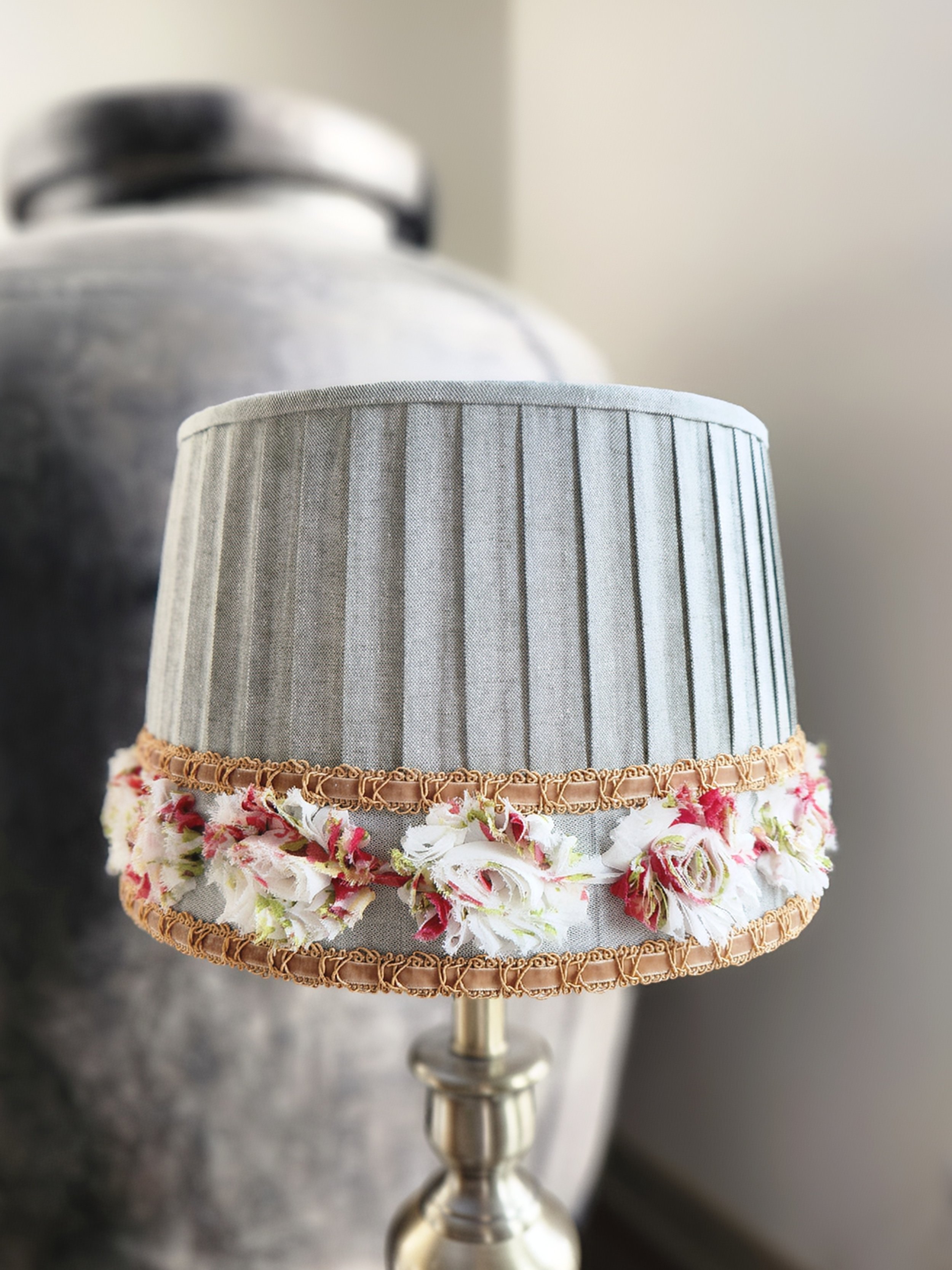 A sage green pleated lampshade adorned with a delicate floral ribbon trim. The ribbon features red, pink and white flowers and is bordered by a gold braid.