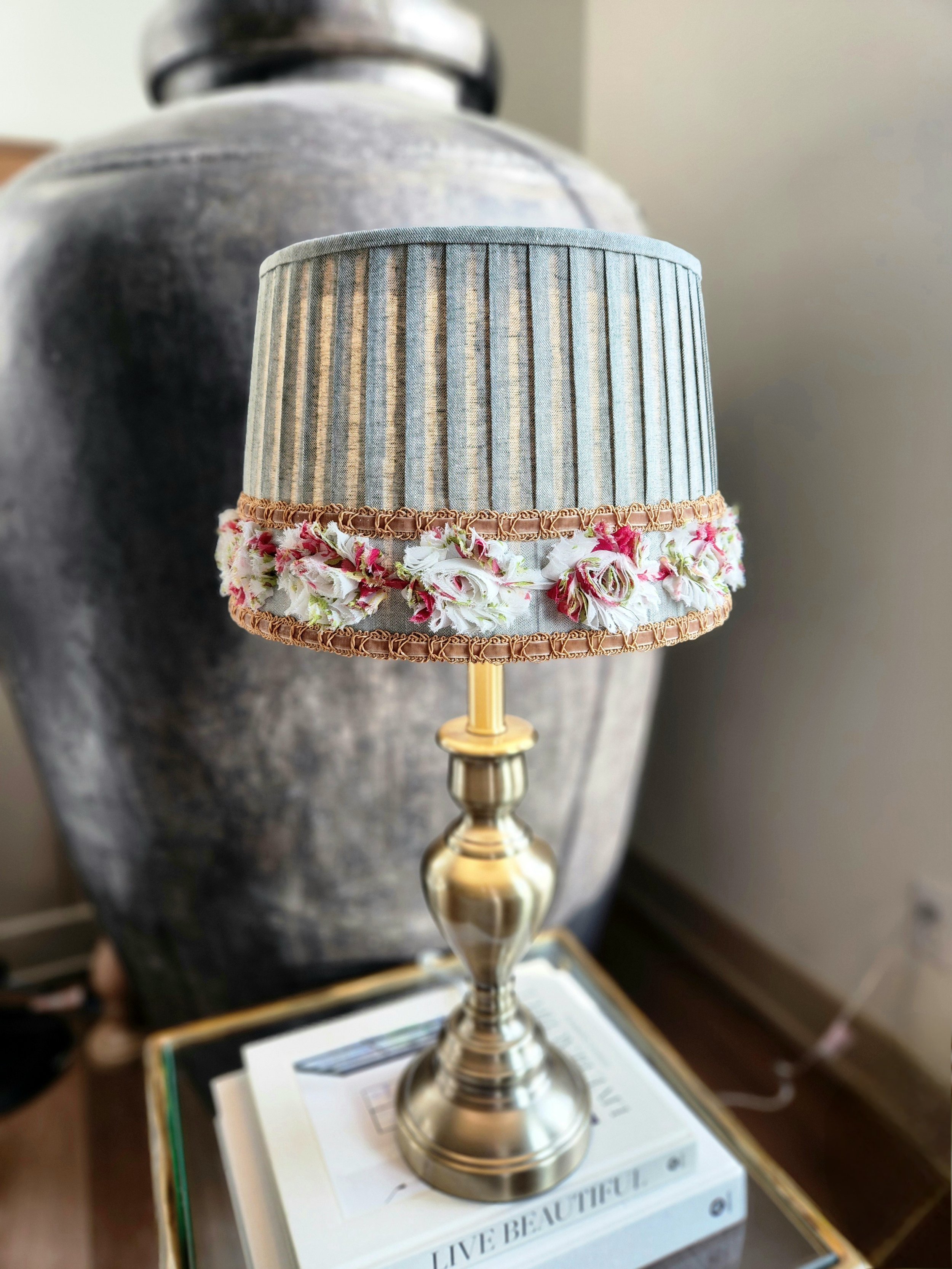 A sage green pleated lampshade adorned with a delicate floral ribbon trim. The ribbon features red, pink and white flowers and is bordered by a gold braid.