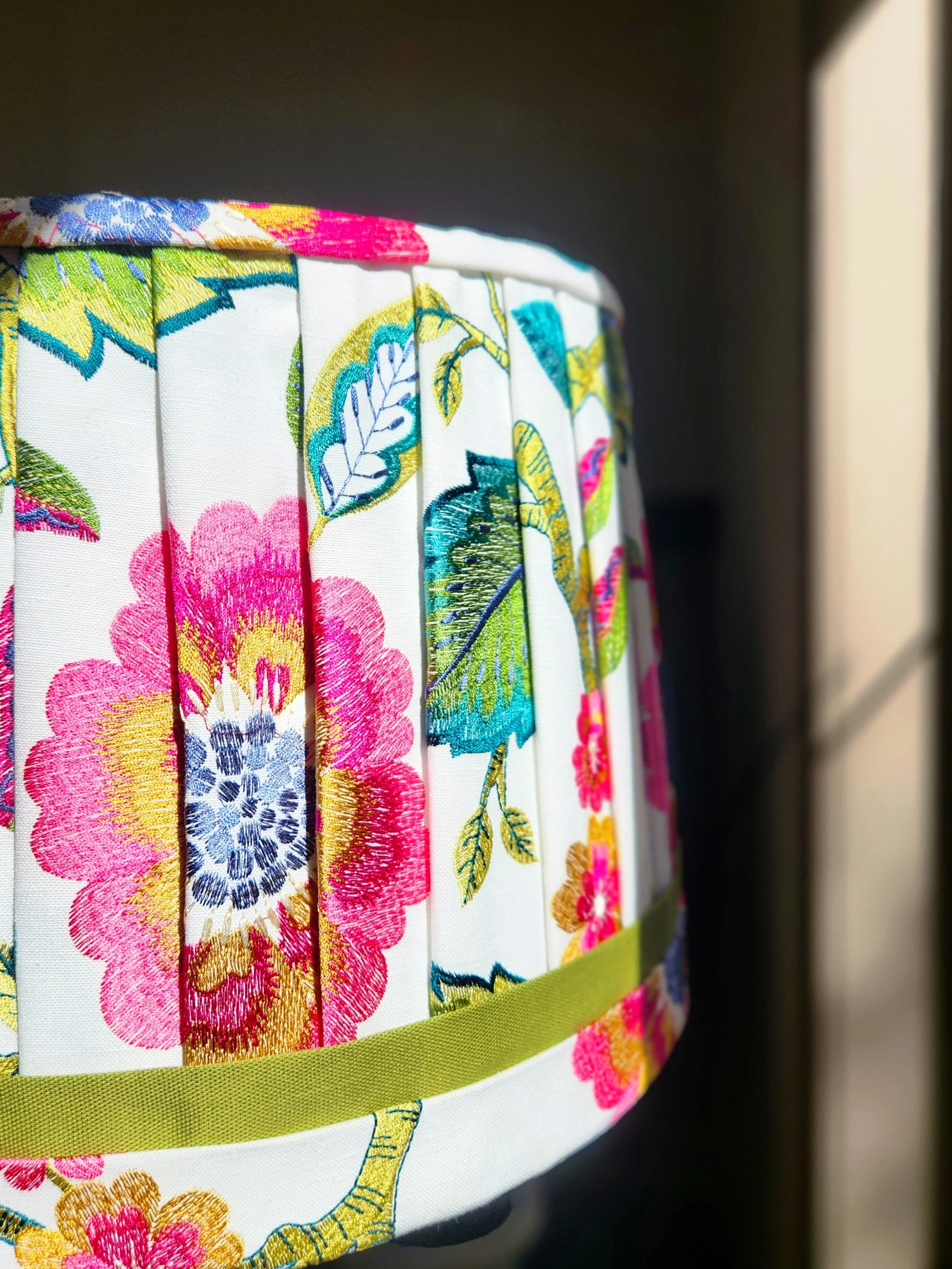 Close-up of a lampshade with vibrant embroidered flowers in pink, blue, and yellow on a white background. The detailed stitching and pleated design are highlighted.