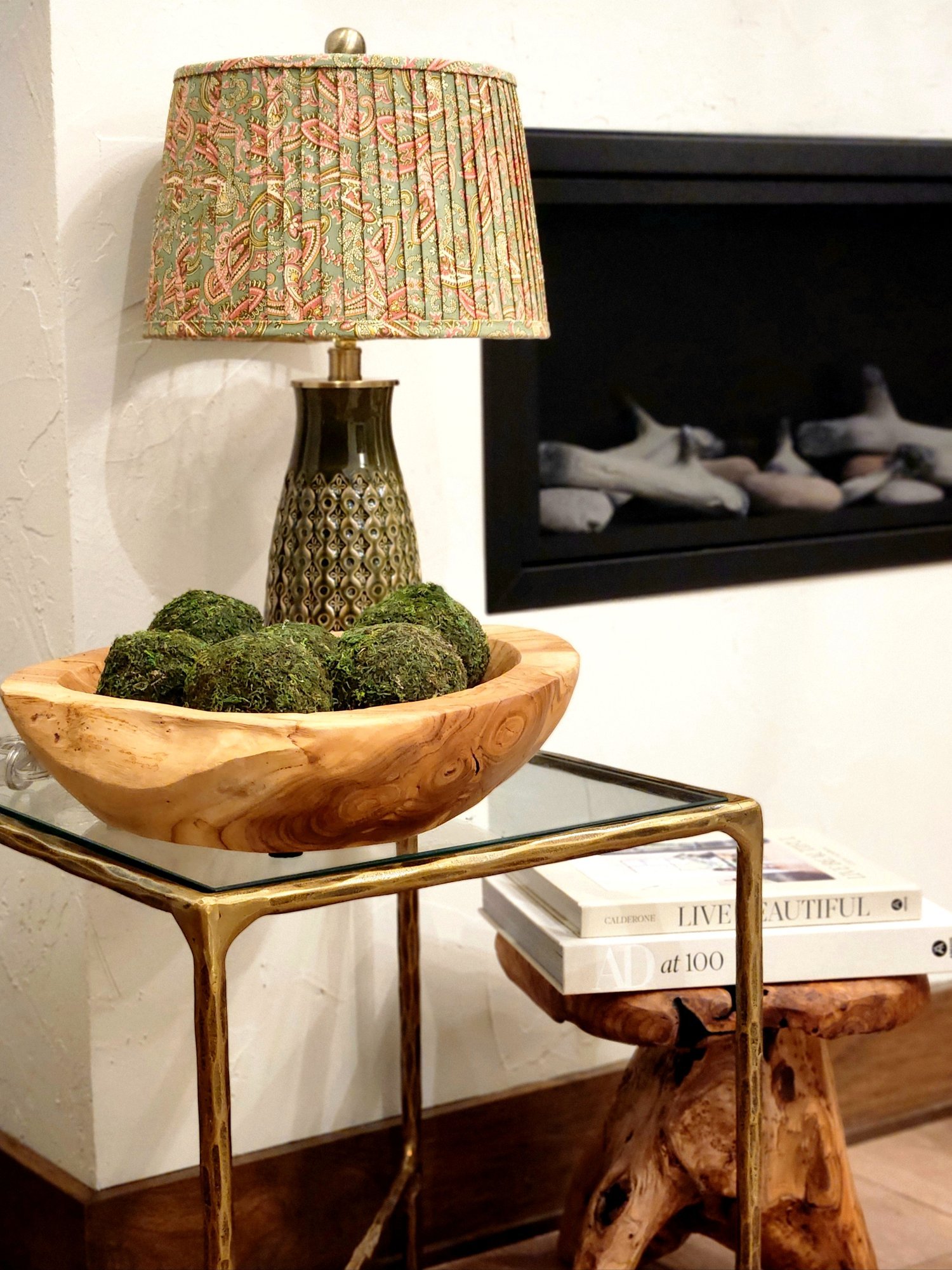 A pink and green paisley patterned lampshade sits on a green ceramic lamp base. They rest on a glass table with a wooden bowl filled with moss and a stack of books underneath.