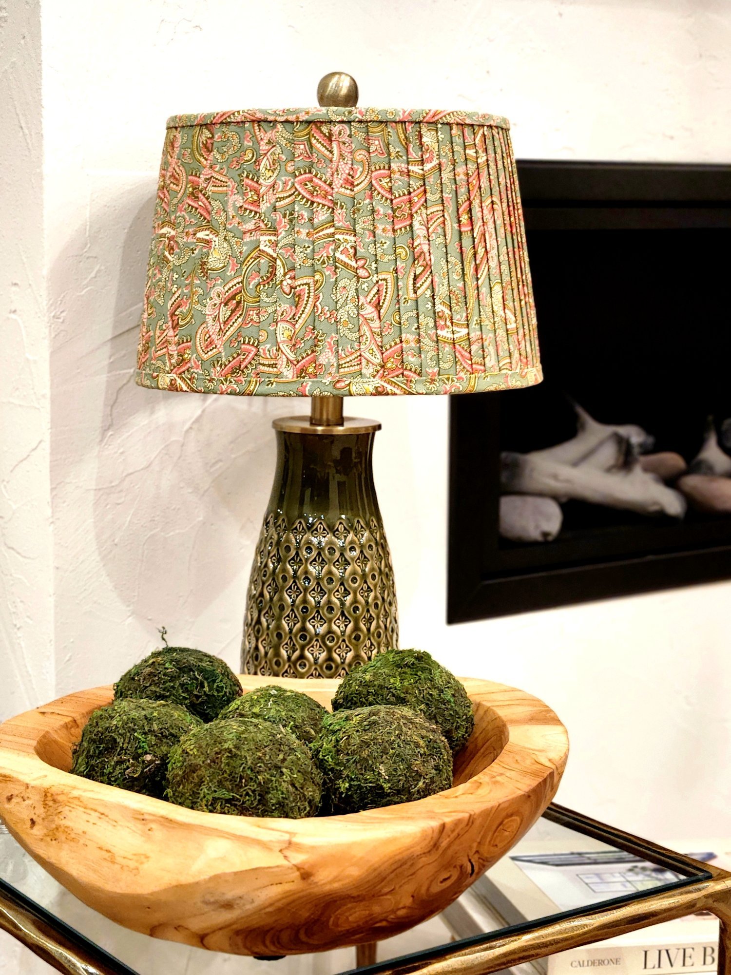 A pink and green paisley patterned lampshade sits on a green ceramic lamp base. They rest on a glass table with a wooden bowl filled with moss and a stack of books underneath.