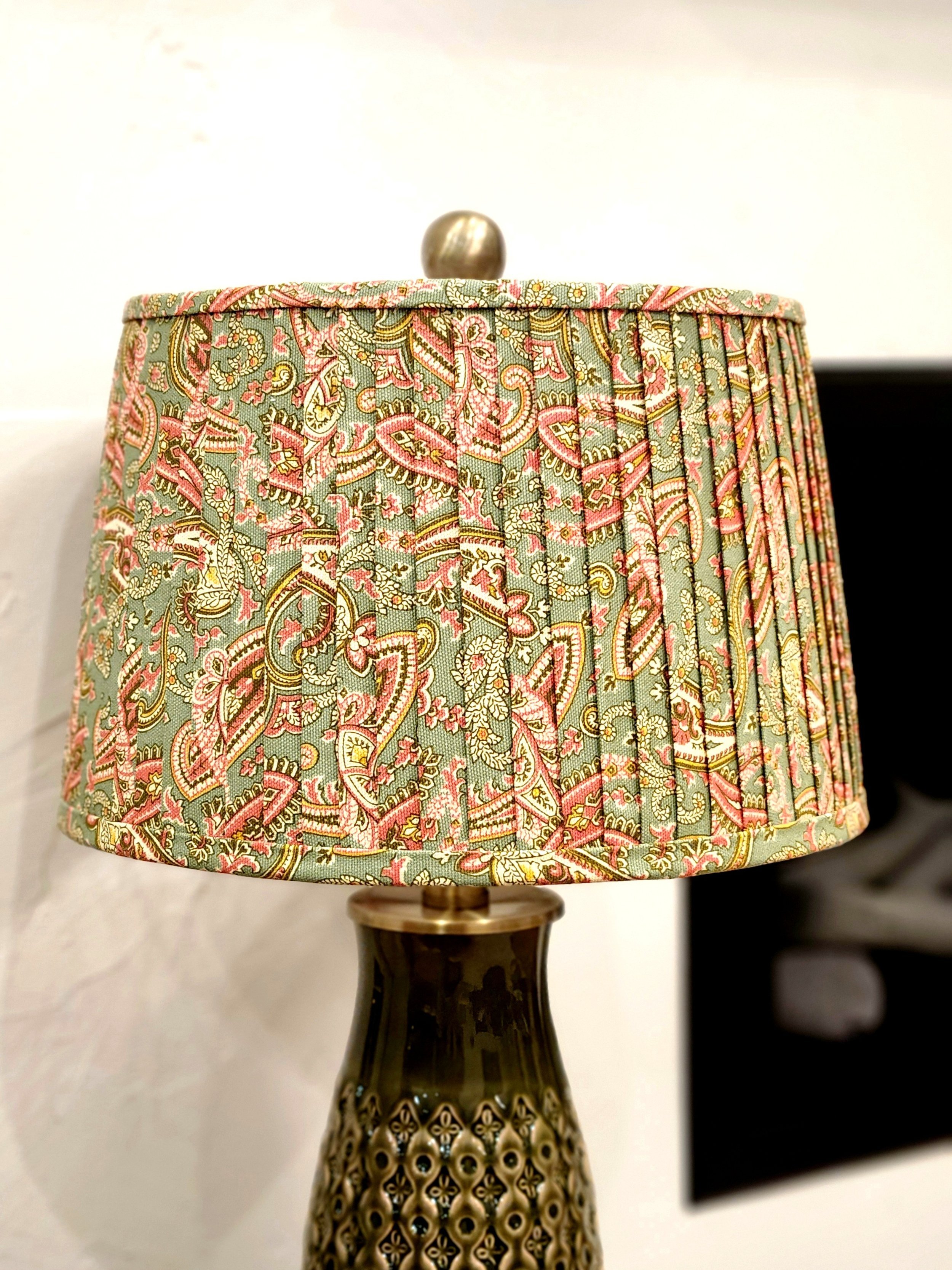 Close-up of a sage green lampshade with a pink and gold paisley pattern. The pleated fabric adds texture and visual interest to the shade.