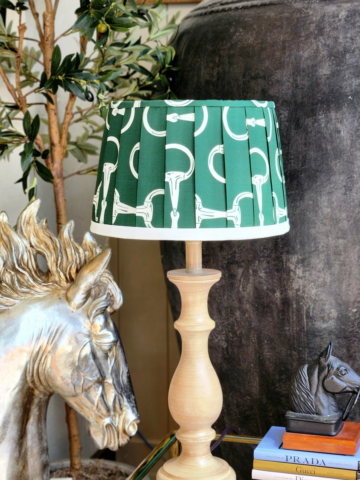 A hunter green lampshade with a repeating pattern of white horse bits. The pleated shade sits on a tan wooden lamp base.