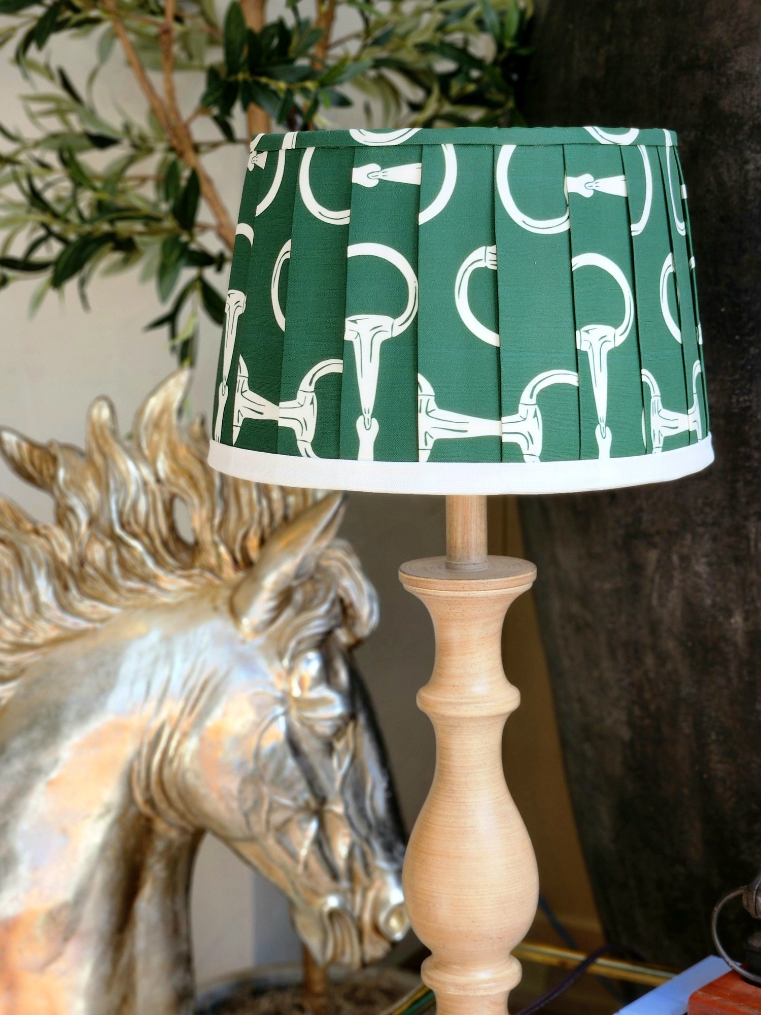 A hunter green lampshade with a repeating pattern of white horse bits. The pleated shade sits on a tan wooden lamp base.