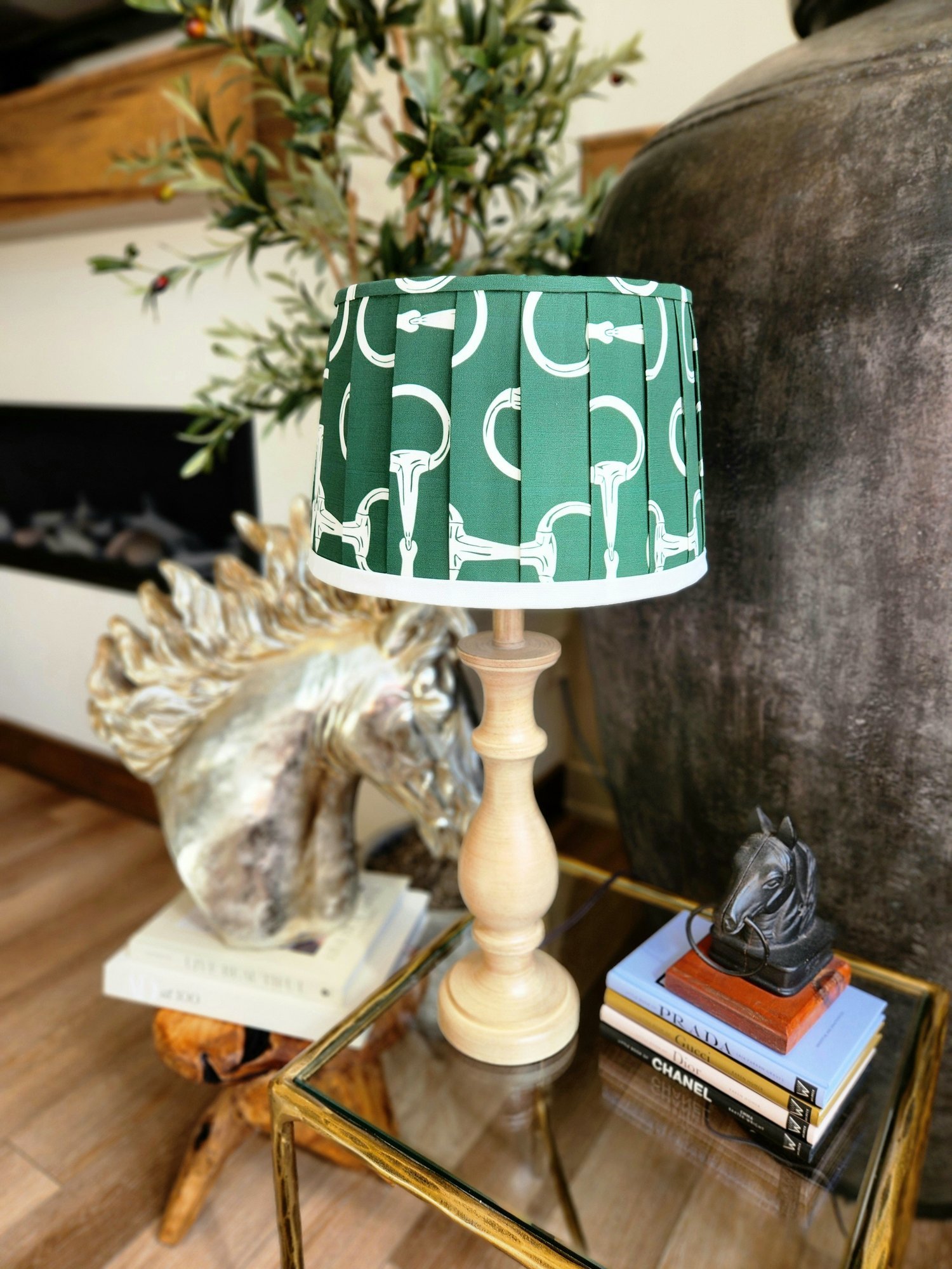 A green lampshade decorated with a pattern of white horse bits sits on a wooden table. A gold horse statue and a stack of books are also visible on the table.