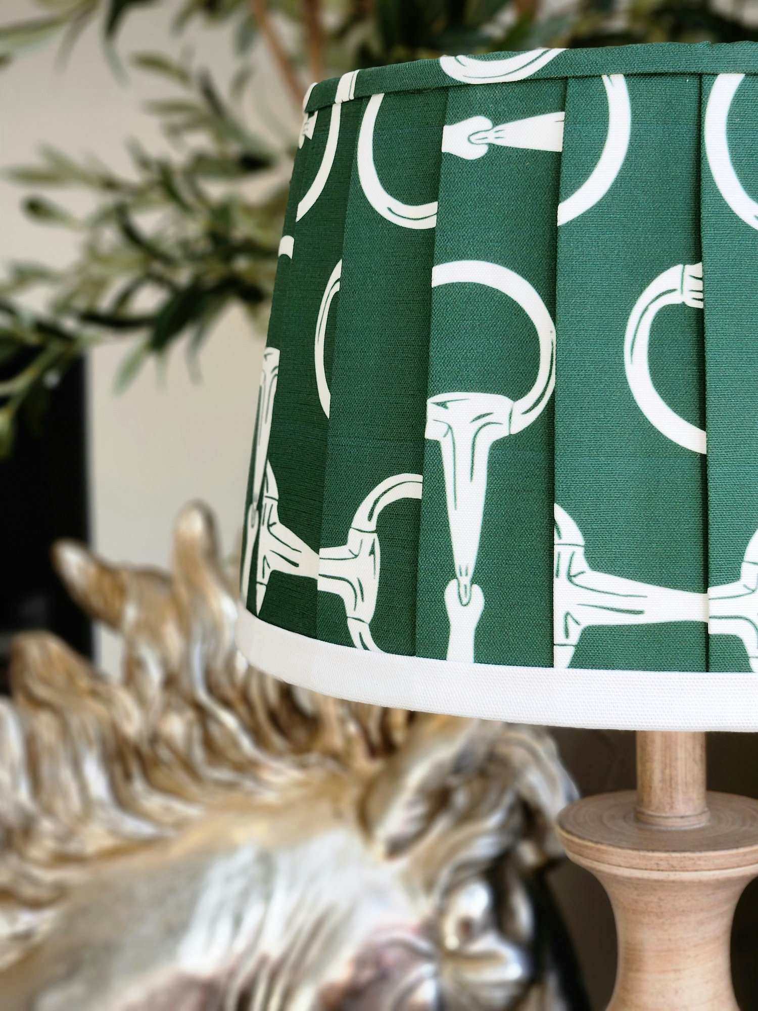 Close-up of a dark green lampshade featuring a repeating pattern of white horse bits. The fabric is pleated, adding texture and dimension.