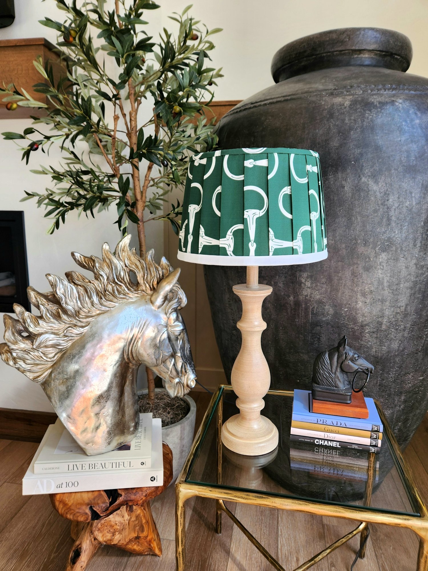 A green lampshade decorated with a pattern of white horse bits sits on a wooden table. A gold horse statue and a stack of books are also visible on the table.