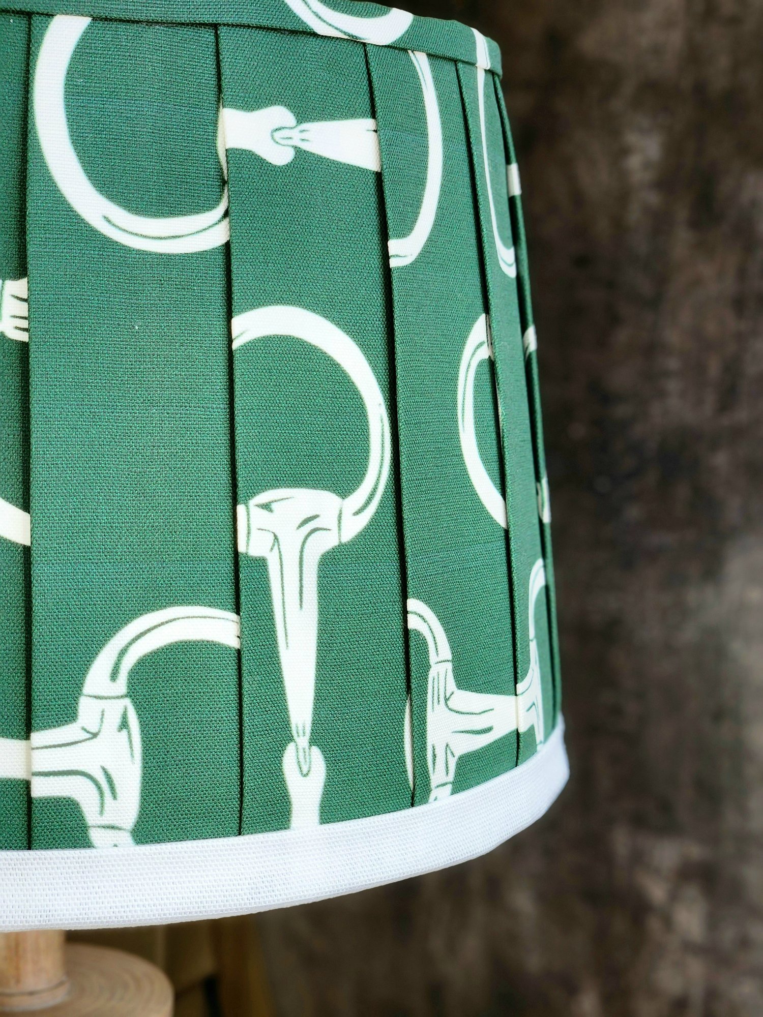 Close-up of a dark green lampshade featuring a repeating pattern of white horse bits. The fabric is pleated, adding texture and dimension.