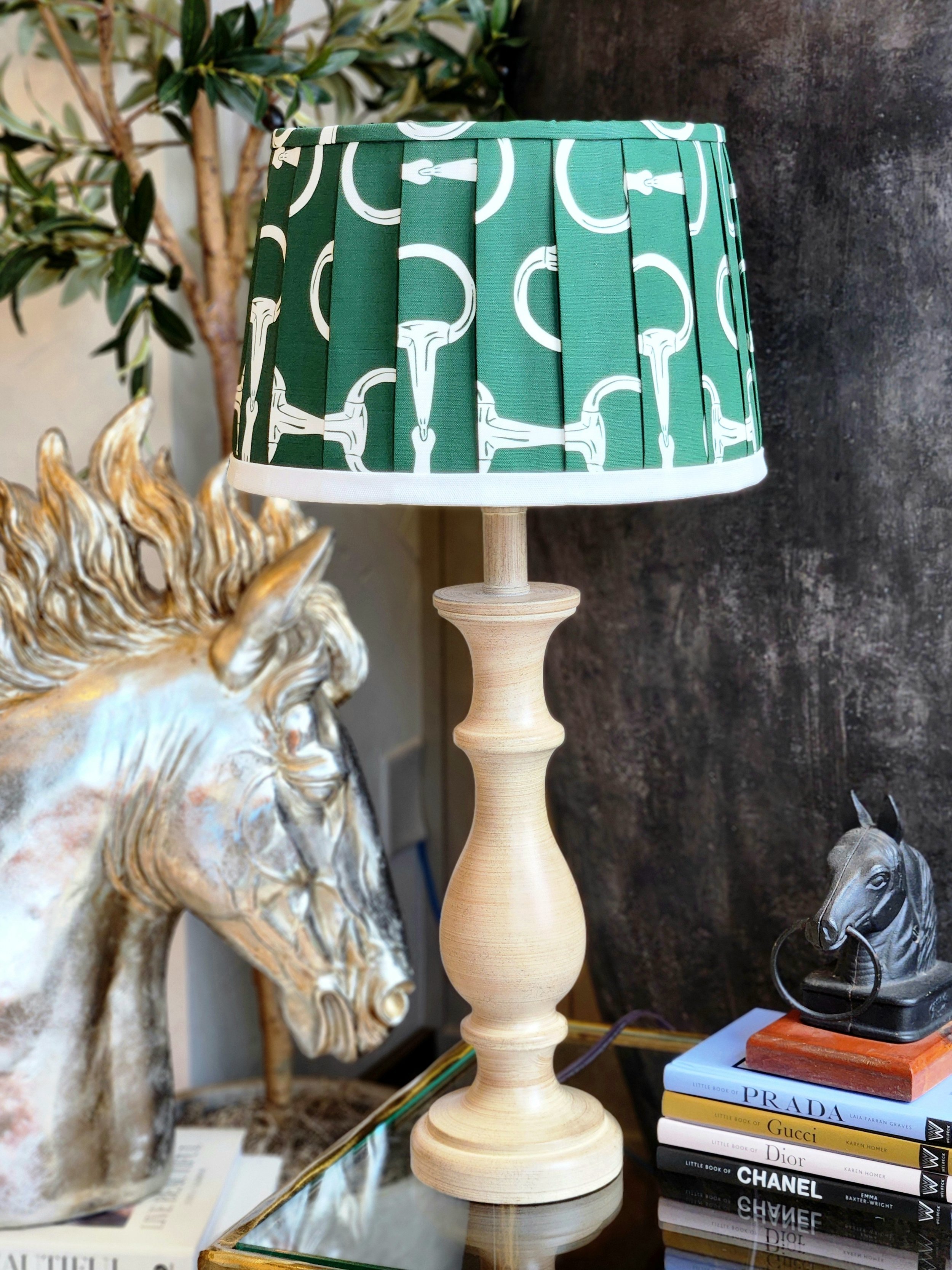 A hunter green lampshade with a repeating pattern of white horse bits. The pleated shade sits on a tan wooden lamp base.