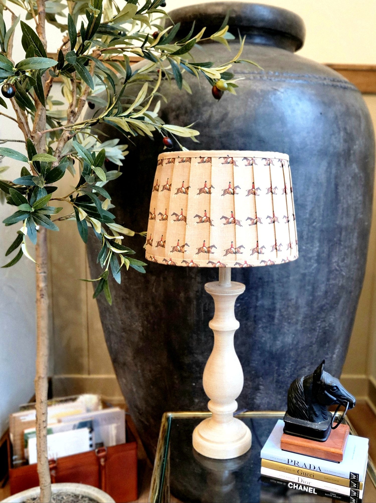 A cream-colored lampshade with a repeating pattern of a jockey riding a horse. The shade is pleated and sits on a light brown wooden lamp base.