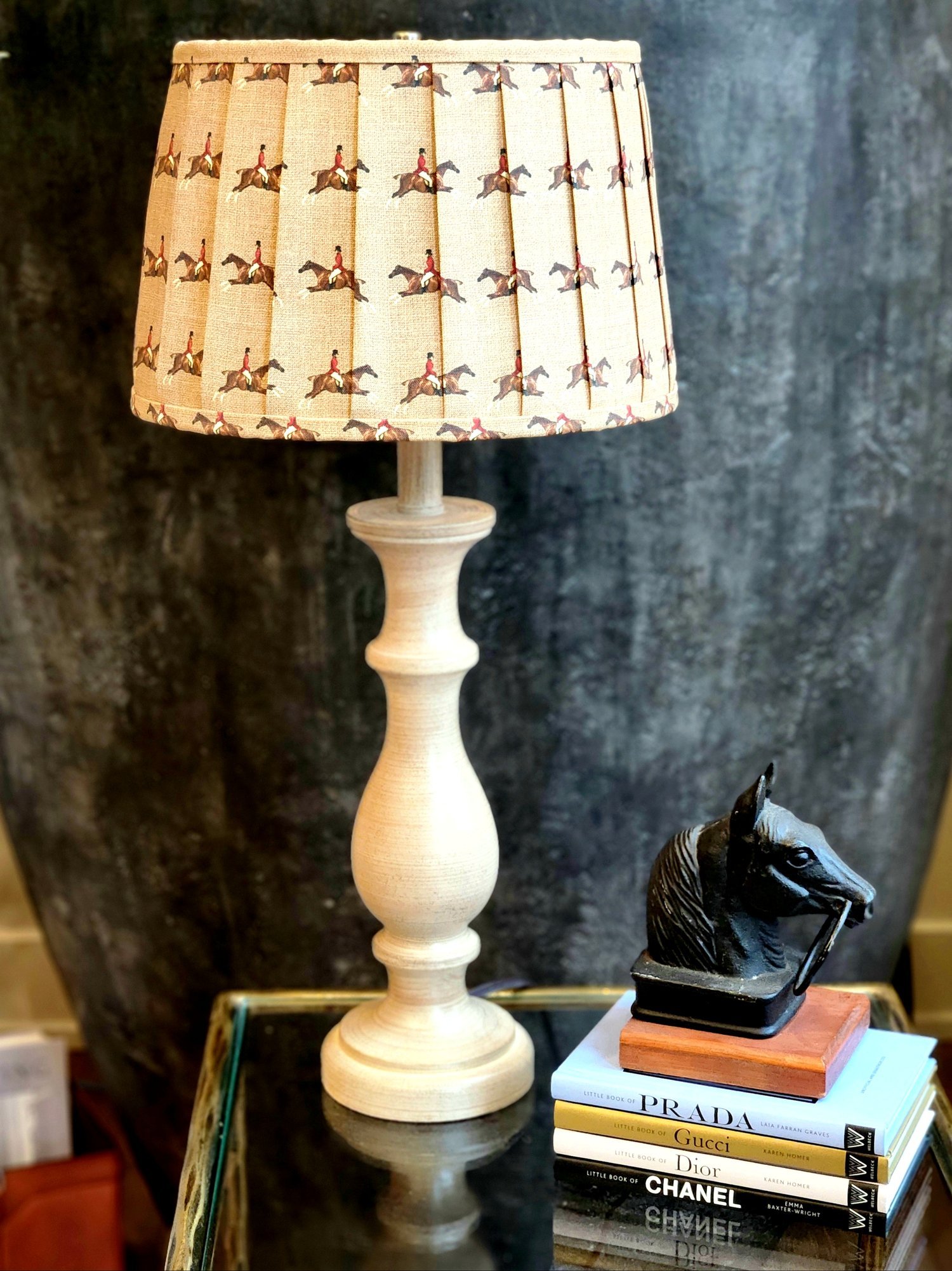 A cream-colored lampshade with a repeating pattern of a jockey riding a horse. The shade is pleated and sits on a light brown wooden lamp base.