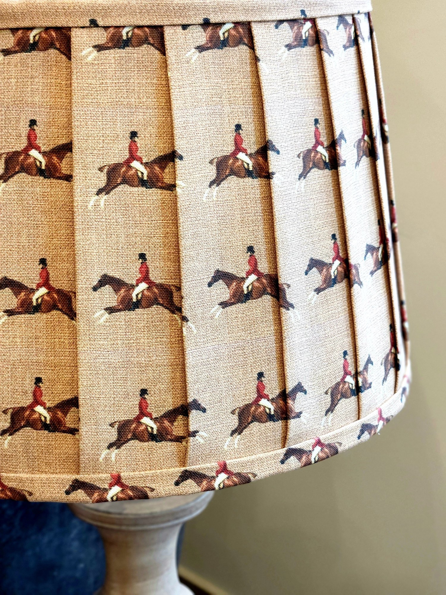 Close-up of a tan lampshade with a repeating pattern of a jockey on a horse. The fabric is pleated, creating vertical lines between each image.