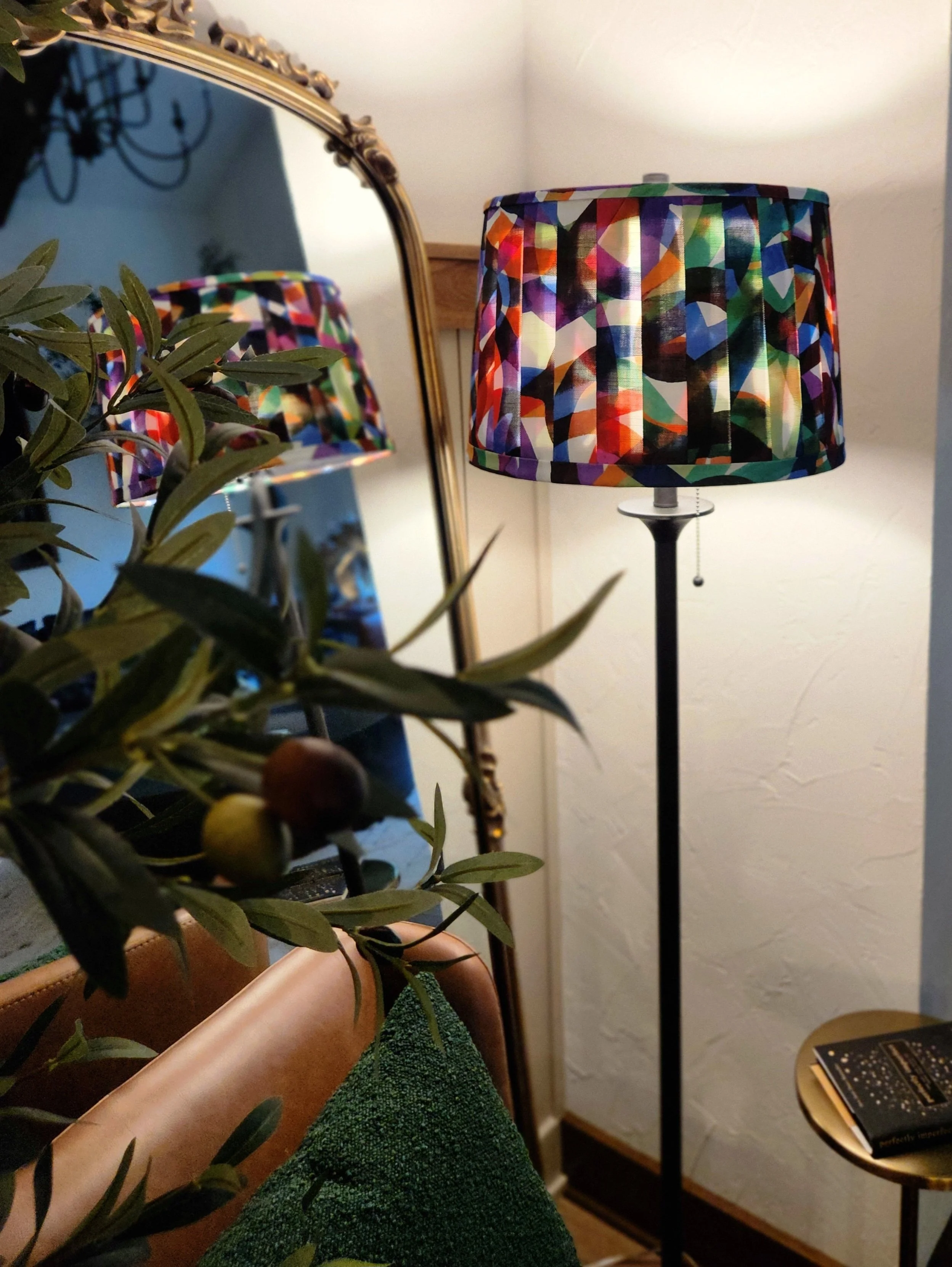 A colorful abstract lampshade, reflected in a mirror. The lamp stands tall with a pleated shade and a black base, adding a vibrant touch to the room.