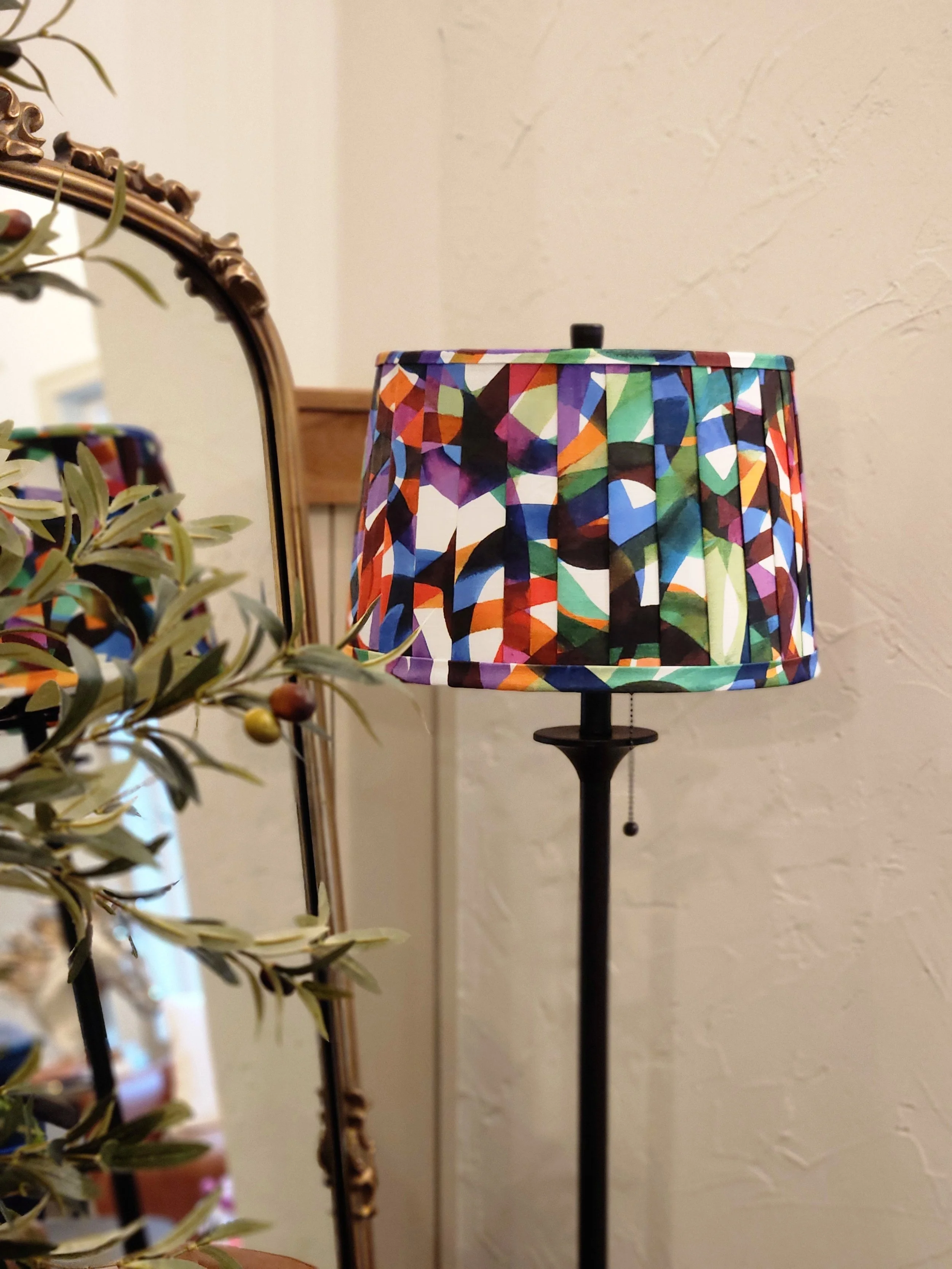 A lamp with a colorful abstract print shade and a pleated design. It stands beside a decorative olive tree and an ornate mirror.