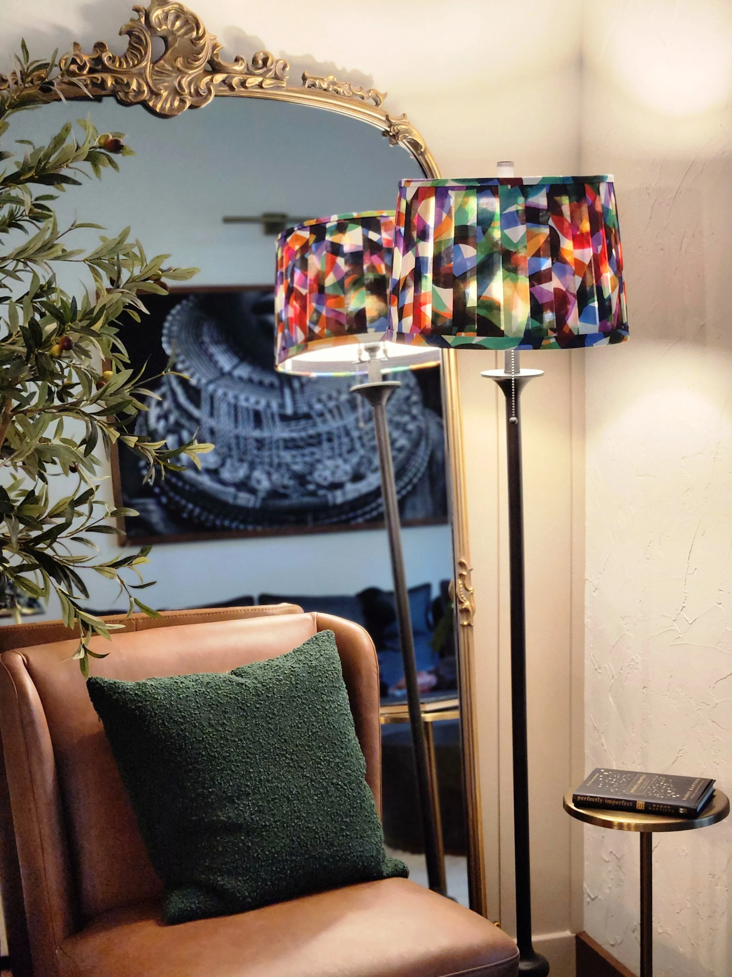 A colorful abstract lampshade, reflected in a mirror. The lamp stands tall with a pleated shade and a black base, adding a vibrant touch to the room.