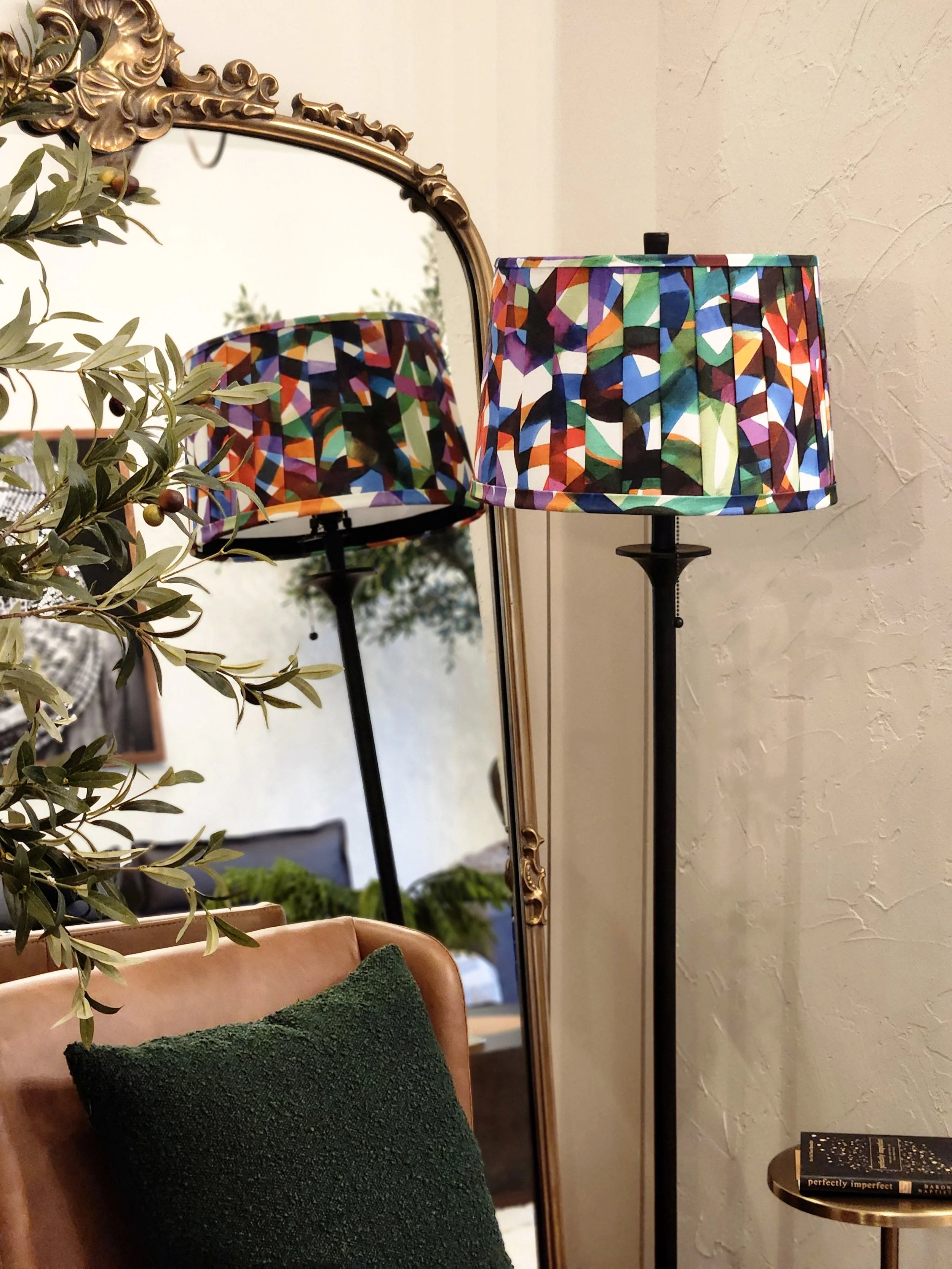 A colorful abstract lampshade, reflected in a mirror. The lamp stands tall with a pleated shade and a black base, adding a vibrant touch to the room.